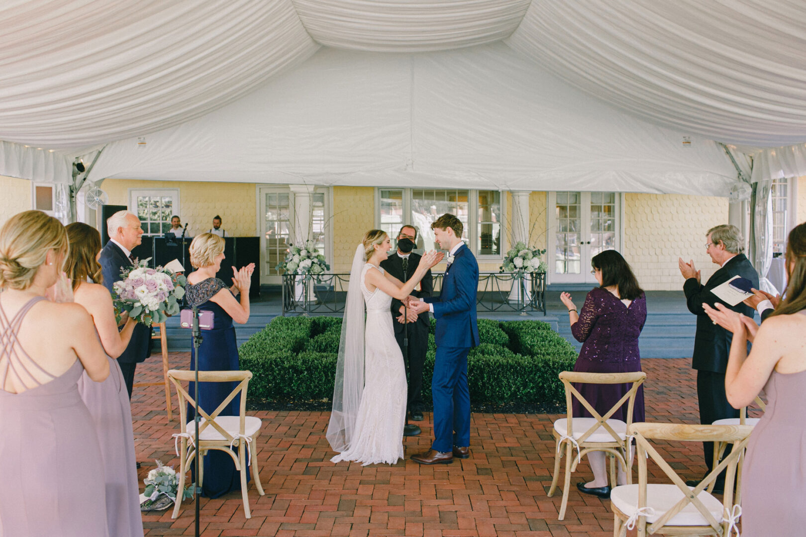 At the Philadelphia Cricket Club wedding, a couple exchanges vows under a white tent during an outdoor ceremony, surrounded by guests clapping and seated on wooden chairs.