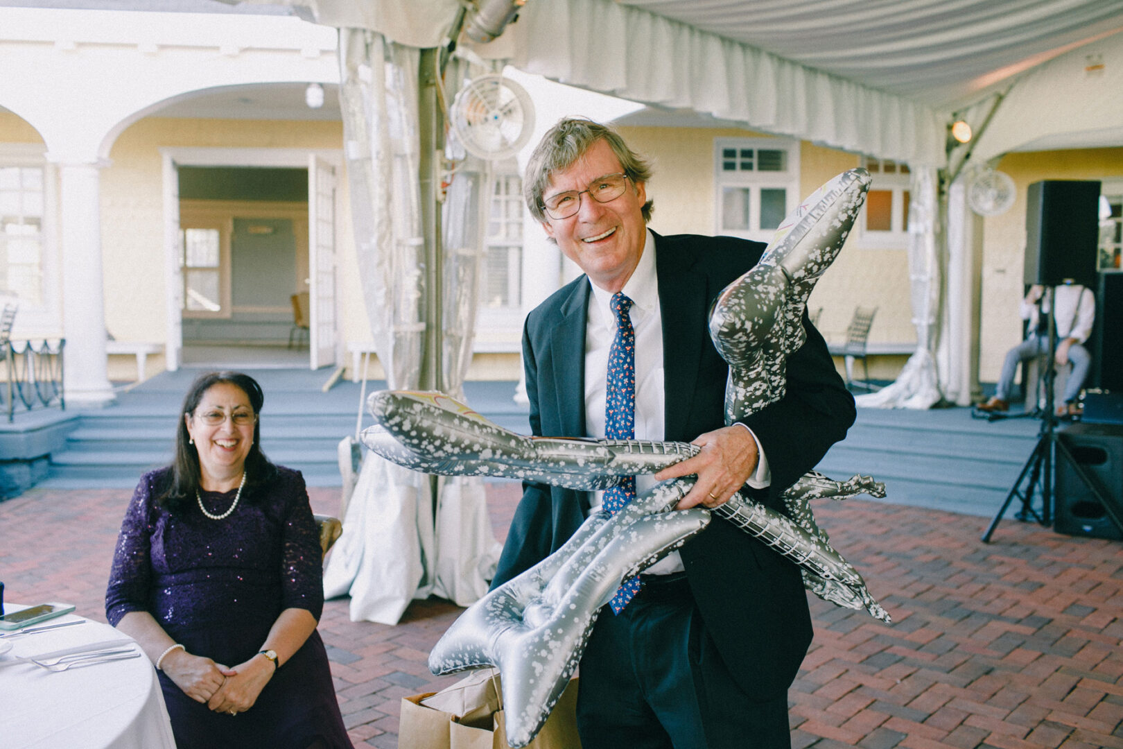 A man in a suit holds silver inflatable objects, smiling, while a seated woman in a purple dress beams. They enjoy the festivities under a white tent at an outdoor event, capturing the delightful ambiance of a Philadelphia Cricket Club wedding.
