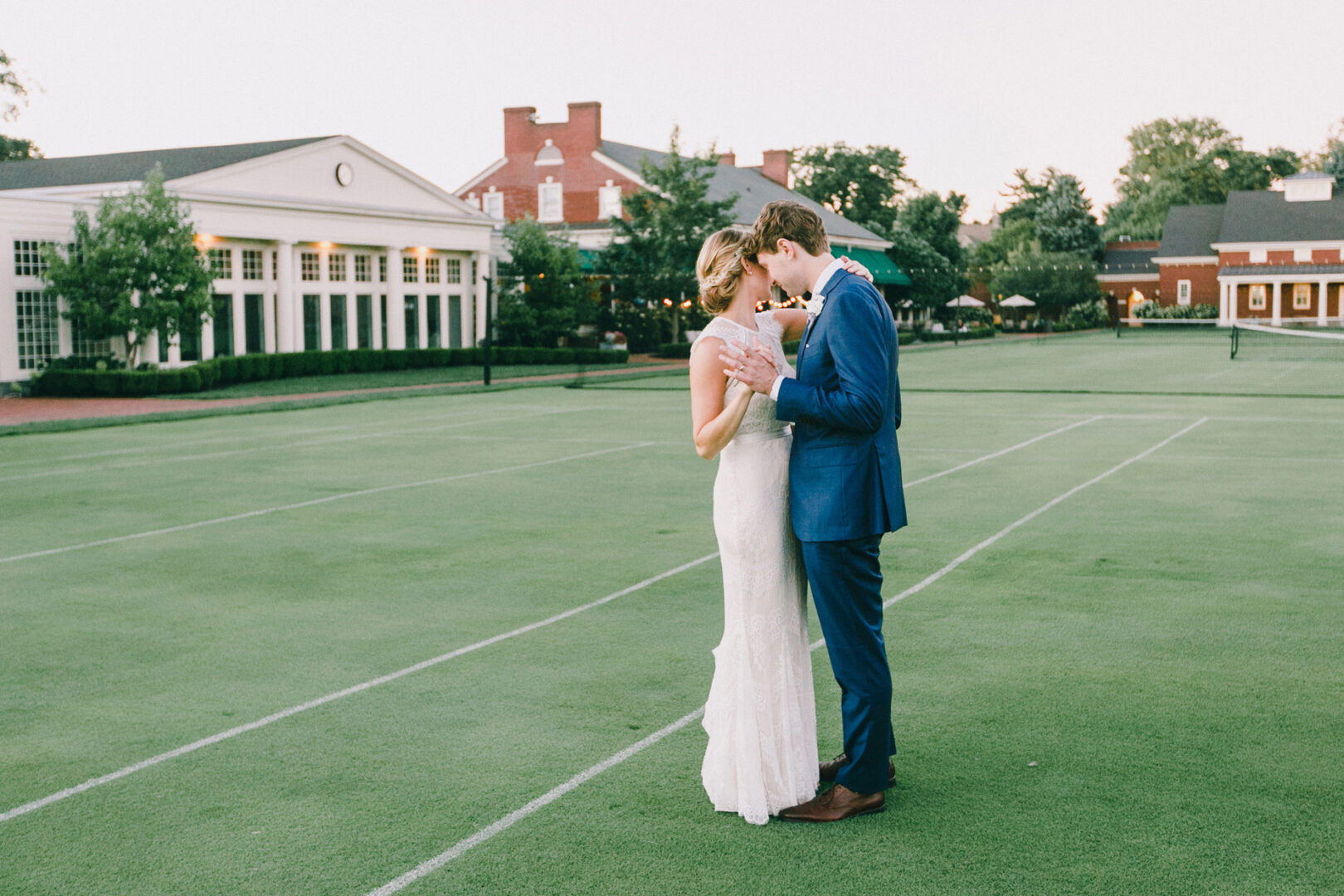 A couple in formal attire dances gracefully on a grassy field, capturing the essence of a Philadelphia Cricket Club Wedding with large buildings as their grand backdrop.