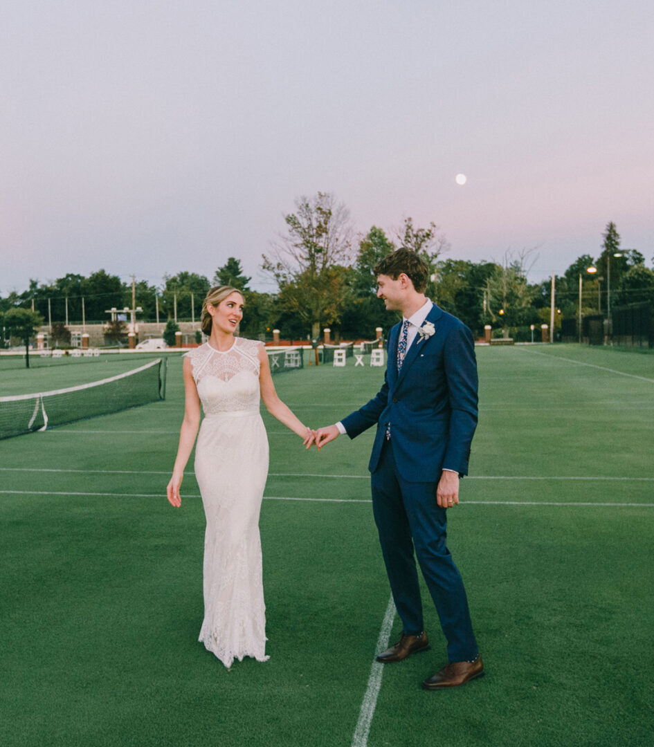 A couple in formal attire holds hands on a tennis court at dusk, embodying the elegance of a Philadelphia Cricket Club wedding, with a single white chair nearby.