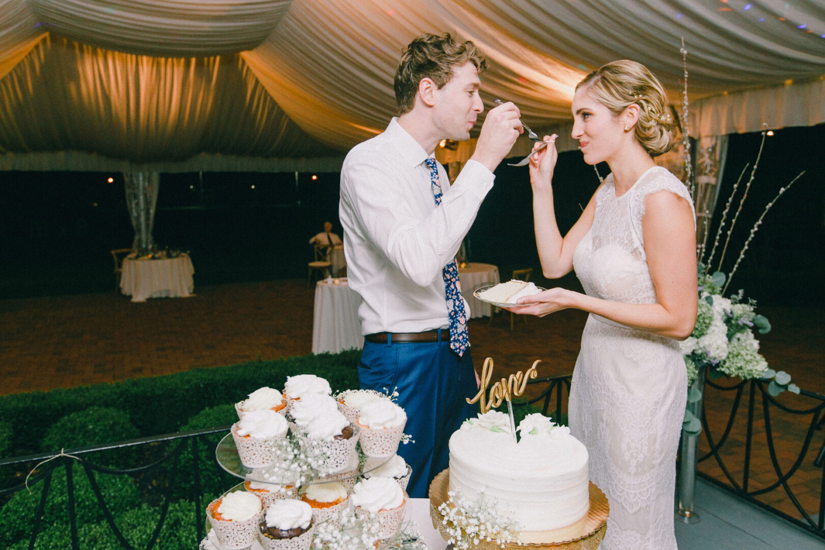 At their Philadelphia Cricket Club wedding, the bride and groom share a slice of cake during the reception. The table is beautifully decorated with cupcakes and a cake topped with a "love" sign.