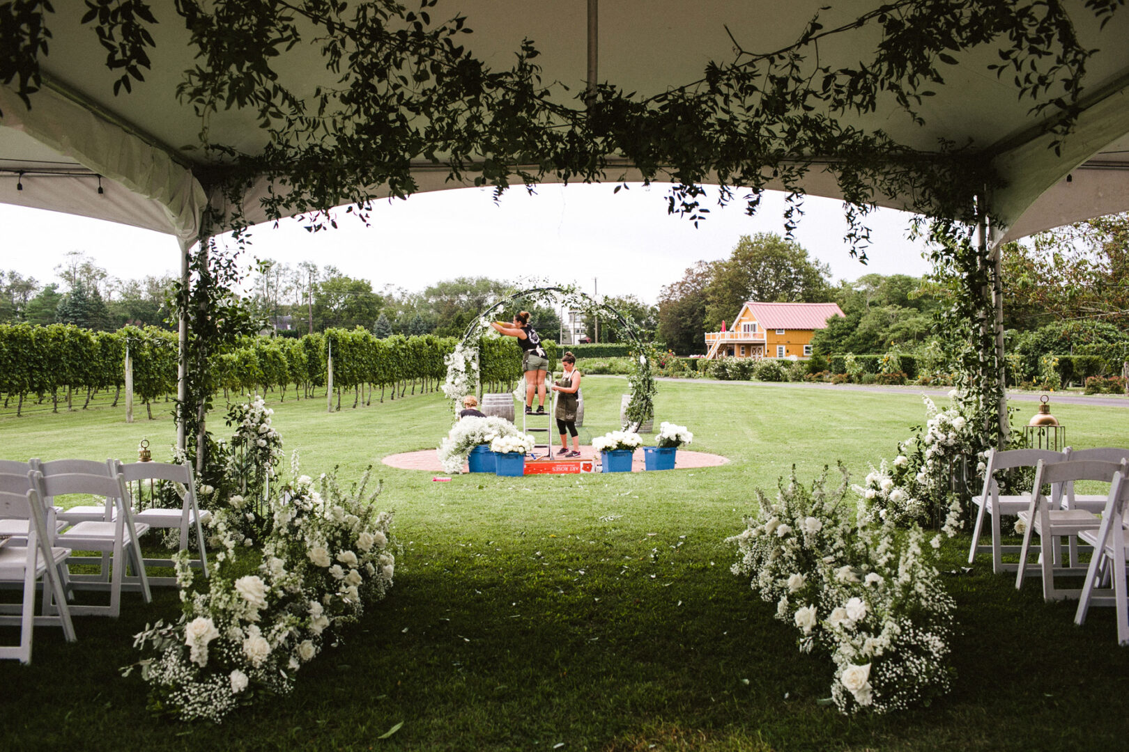 A stunning Willow Creek Winery wedding unfolds with a ceremony setup under a tent adorned with white flowers and lush greenery. Two people gracefully arrange decorations, perfecting the enchanting ambiance of this grassy outdoor venue.