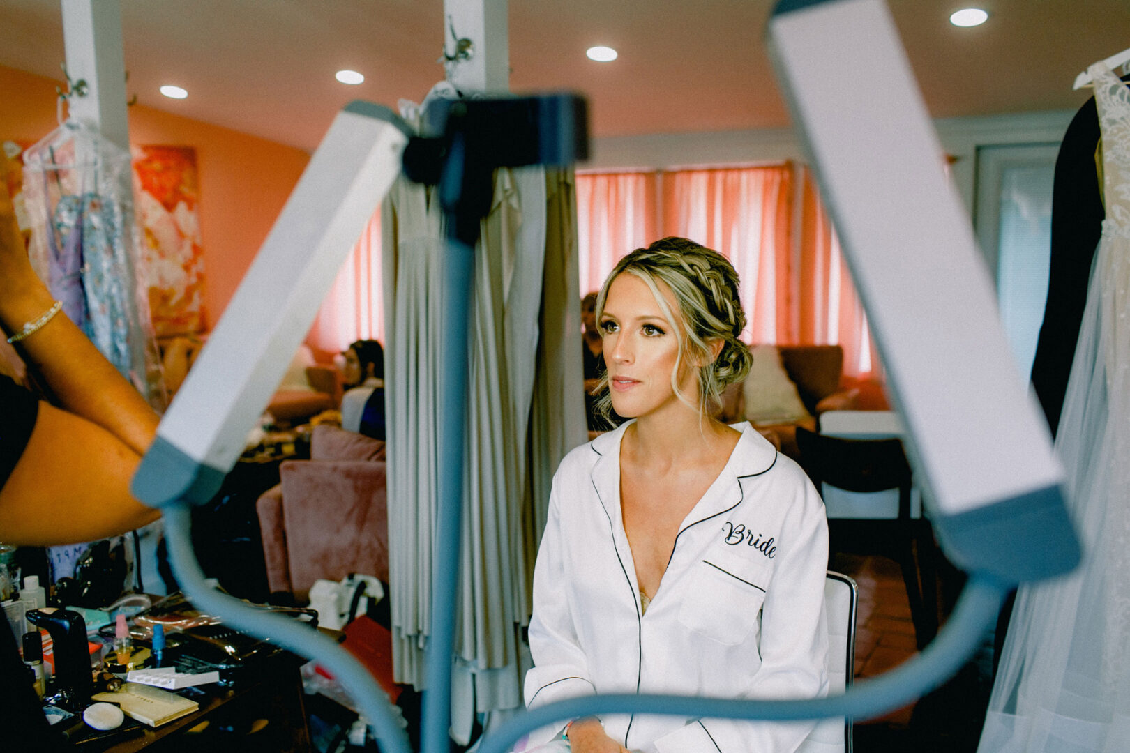 A woman in a white robe, with "Bride" embroidered, sits in front of mirrors and light panels at her Willow Creek Winery wedding, getting her hair styled in a room filled with clothing and makeup supplies.