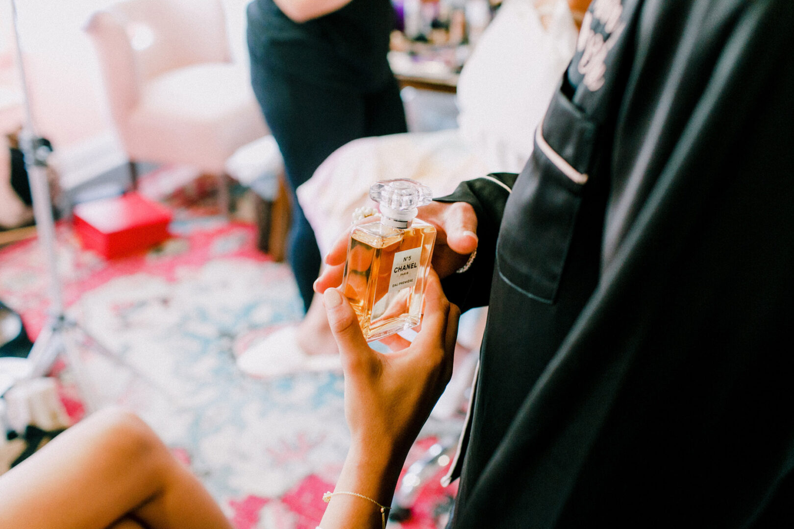 A person holds a bottle of Chanel perfume indoors, evoking the elegance of a Willow Creek Winery wedding. A patterned rug and a chair add warmth and charm to the background.