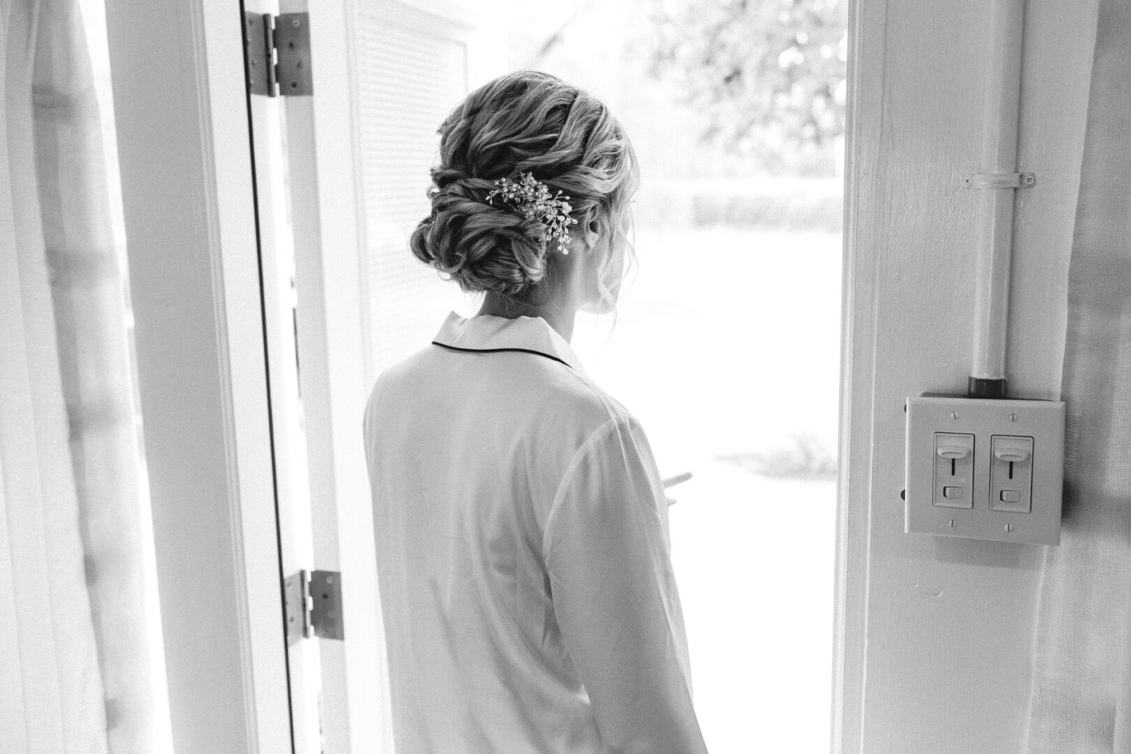 A woman with styled hair and a floral accessory stands by an open door at Willow Creek Winery, gazing outside. Black and white image.