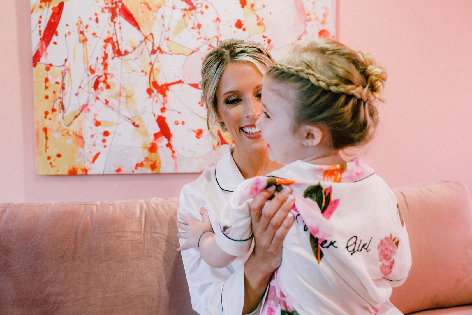 A woman smiles at a young child wearing floral pajamas. They sit on a pink sofa, with an abstract painting reminiscent of the vibrant art found at Willow Creek Winery weddings in the background.
