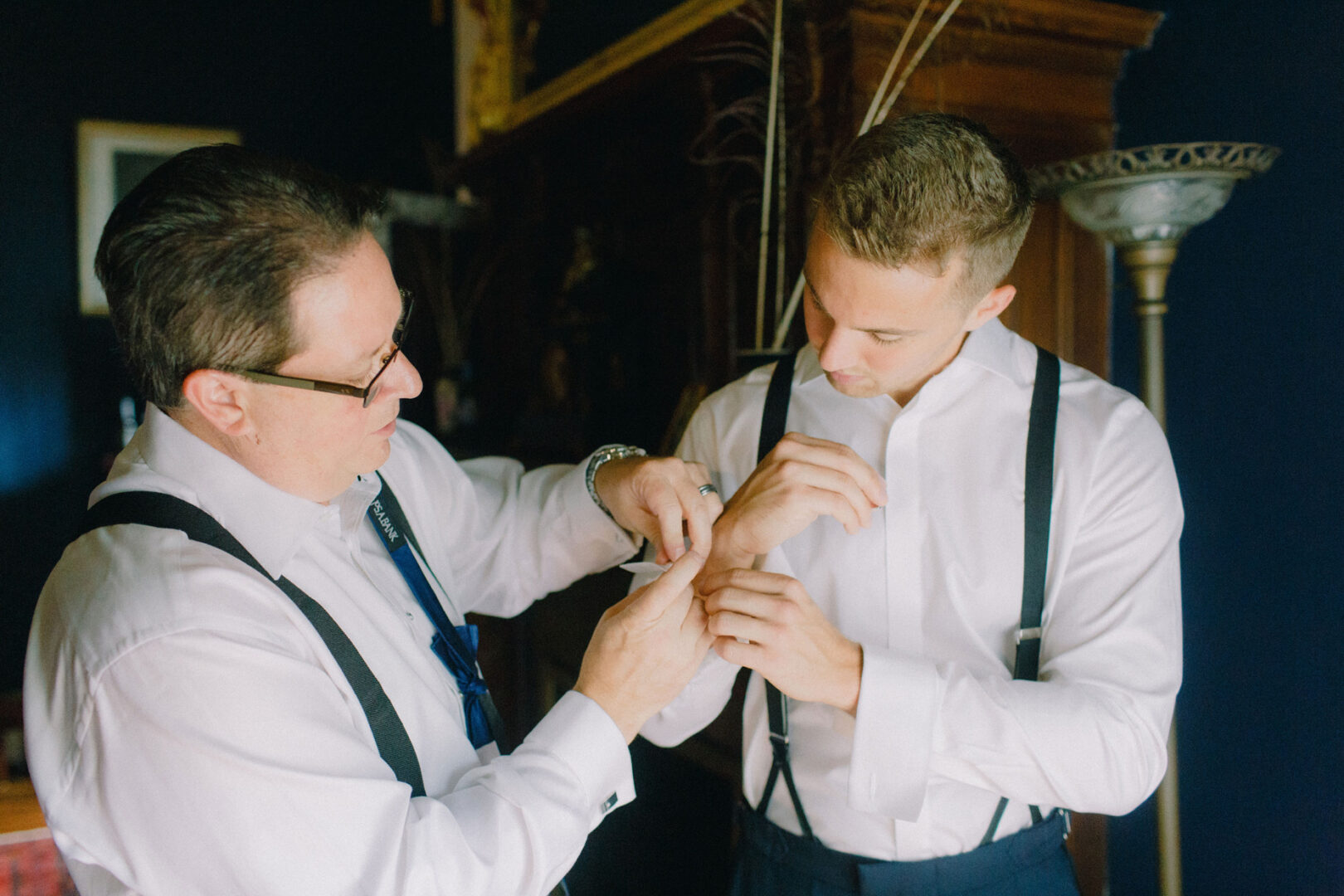 Two men in formal attire adjust cufflinks, one assisting the other, as they prepare for a Willow Creek Winery wedding. The room's dark walls and a softly glowing floor lamp create an intimate atmosphere for this special occasion.