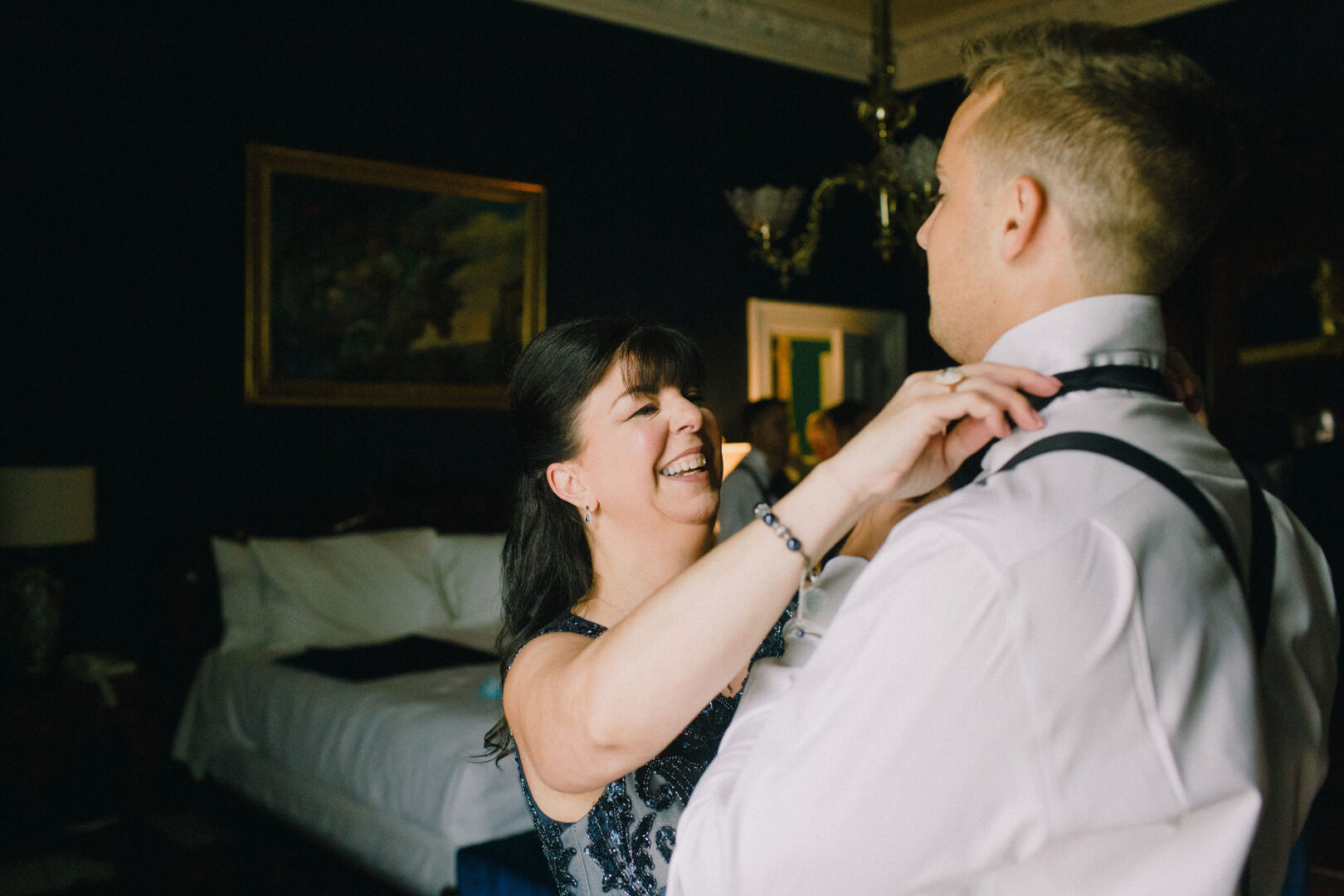 In a dimly lit room with elegant decor, a woman helps a man adjust his bow tie, setting the scene for an unforgettable Willow Creek Winery wedding.