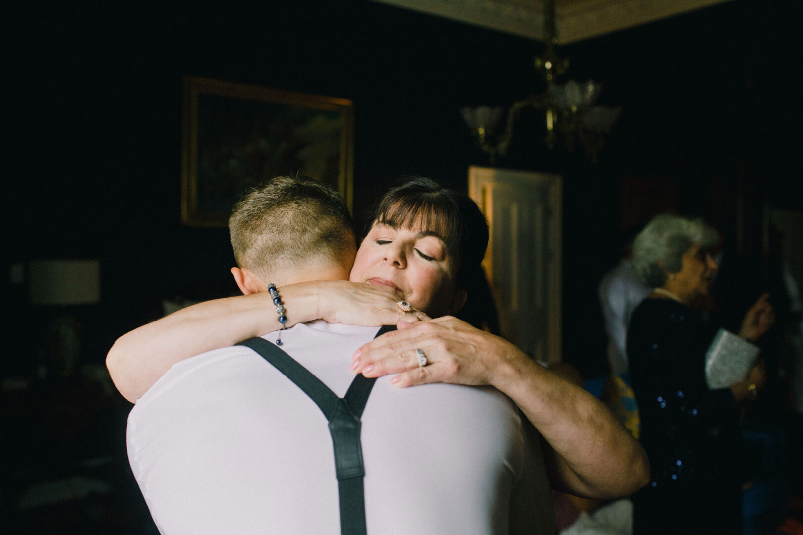 In a dimly lit room at Willow Creek Winery, a woman embraces a man in a white shirt with suspenders. Another person lingers in the background, adding to the intimate ambiance of the wedding celebration.
