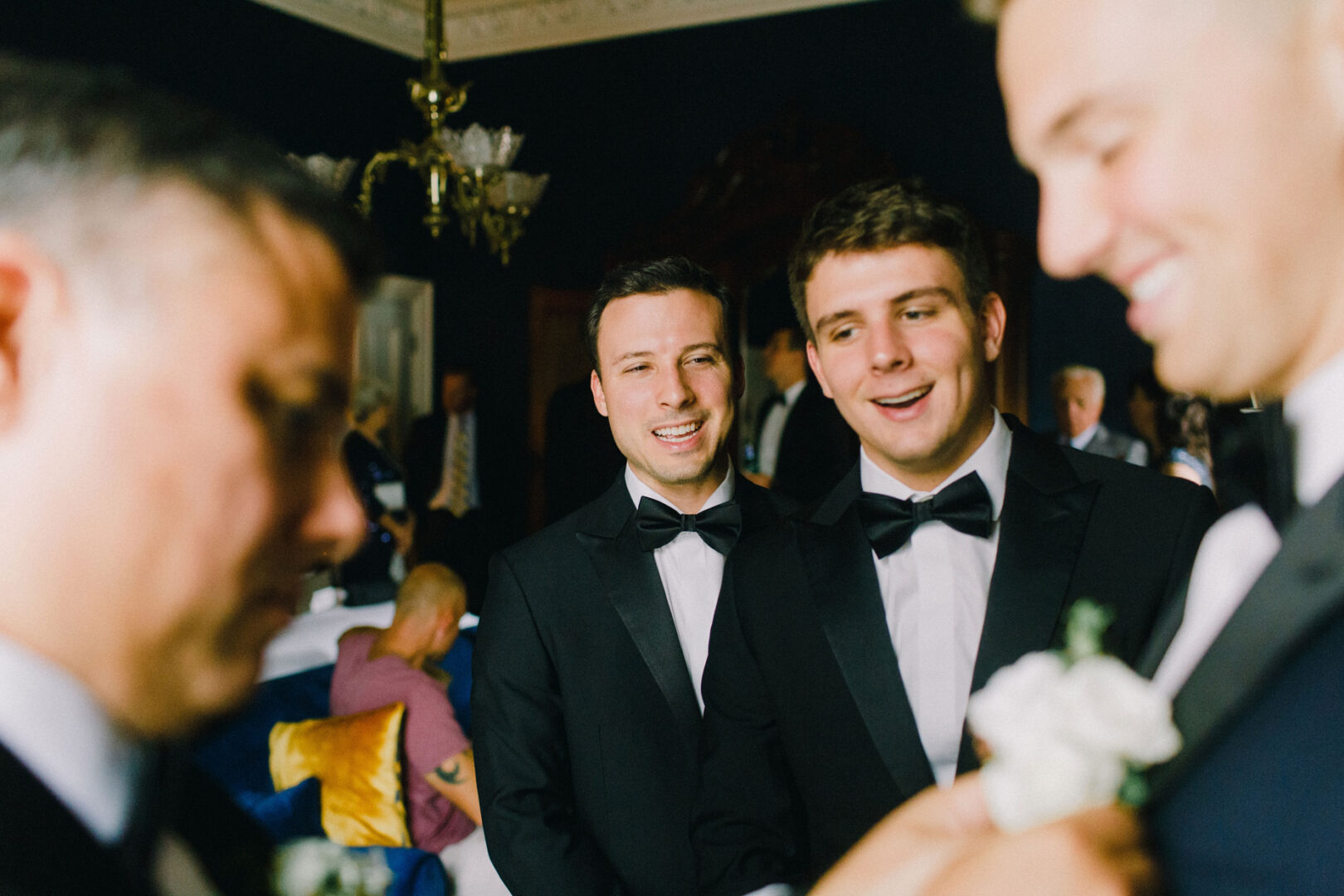 A group of men in suits and bow ties stand together, smiling and talking in a well-lit room with a chandelier, celebrating the Willow Creek Winery wedding.