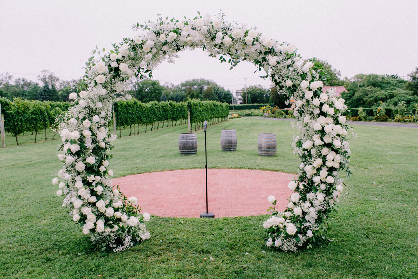 A white floral archway graces the green lawn at Willow Creek Winery, with three wooden barrels in the background. Nearby trees sway gently under a cloudy sky, setting a picturesque scene for a perfect wedding day.