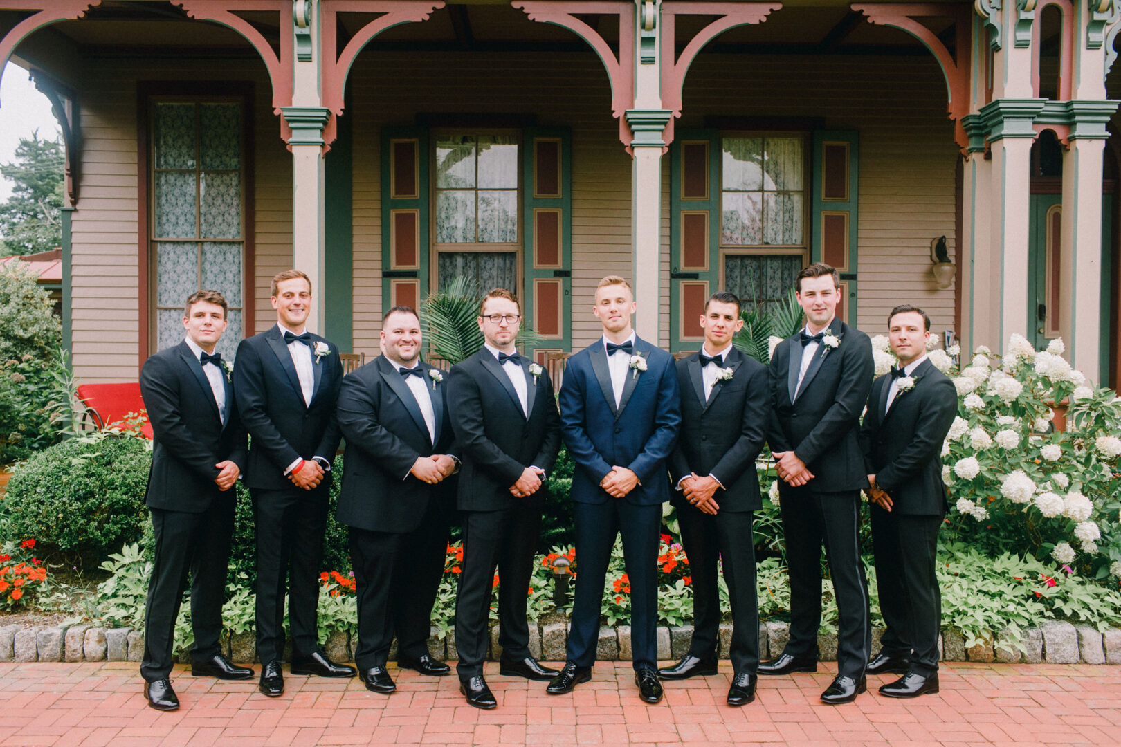 Eight groomsmen in tuxedos stand in front of a Victorian-style house with detailed woodwork and a garden, setting the perfect scene for a Willow Creek Winery wedding.