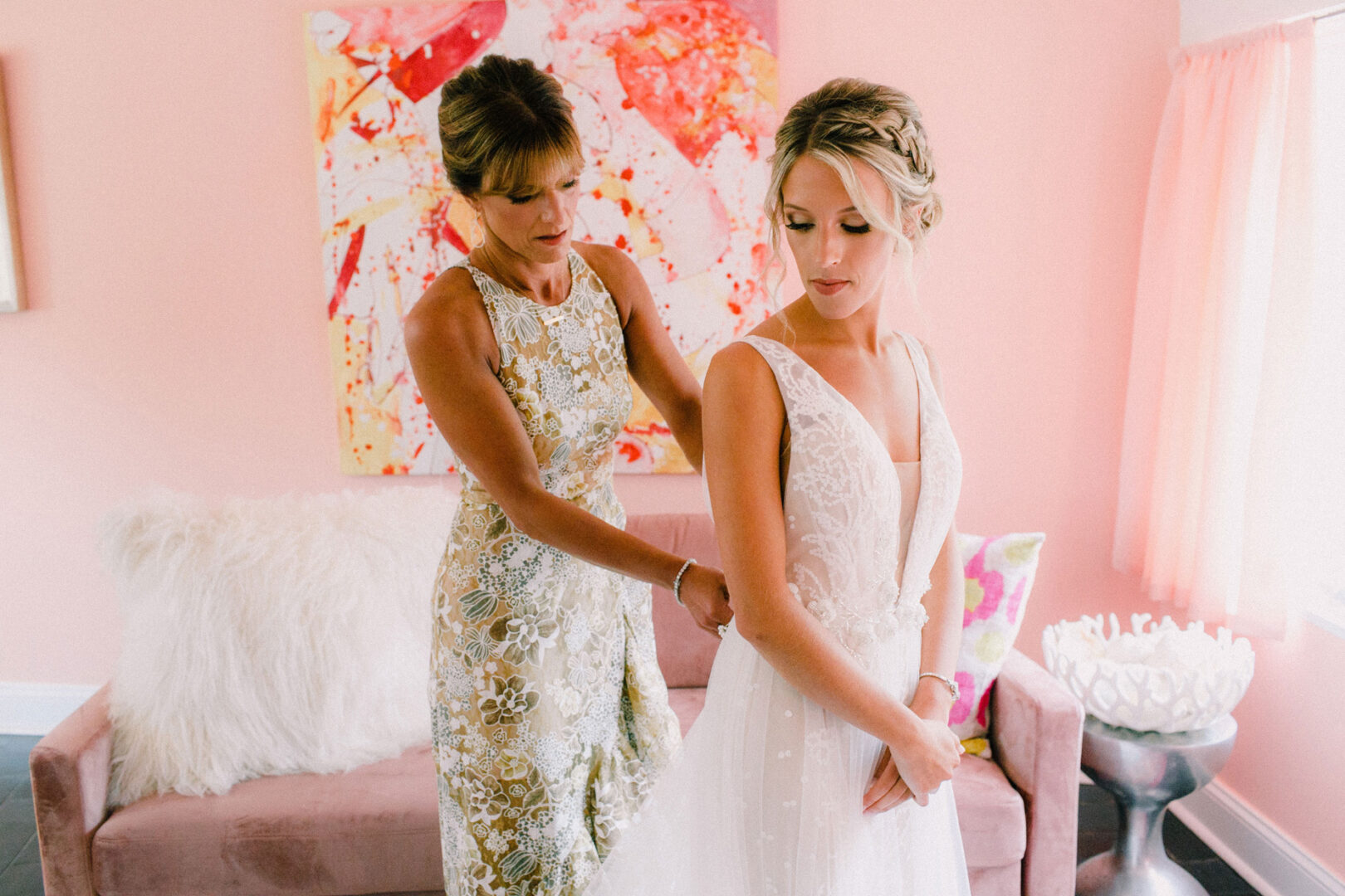 In a room with pink walls and a colorful painting, a woman assists another in adjusting her wedding dress, adding the perfect touch for the upcoming Willow Creek Winery wedding.