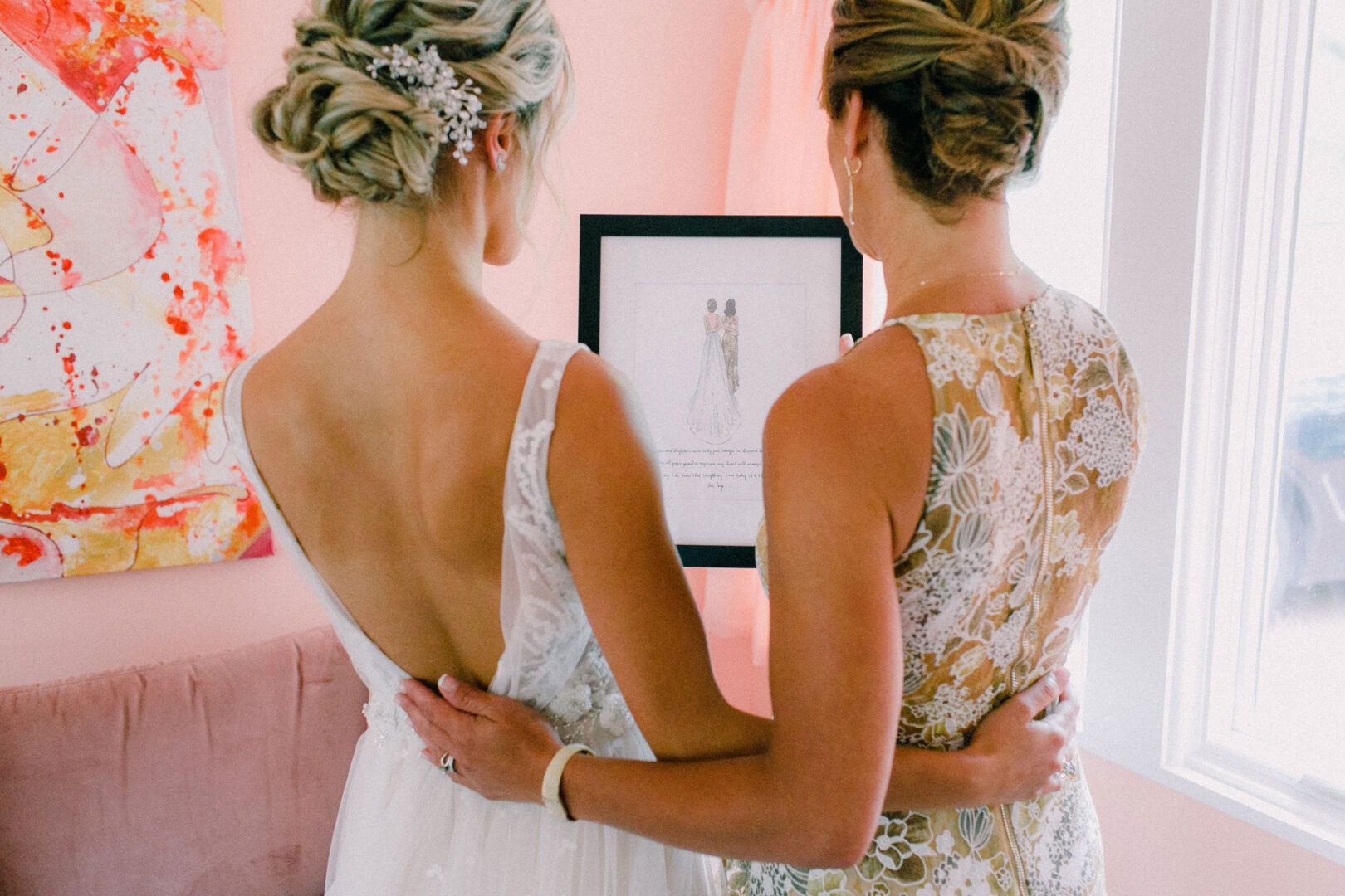 Two women in formal dresses, viewed from the back, stand together at a Willow Creek Winery wedding, admiring a framed picture. One woman has an updo adorned with decorative hair accessories, perfectly capturing the elegance of the occasion.