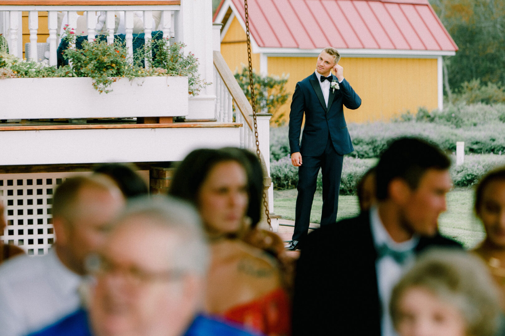 A person in a suit stands on the porch of Willow Creek Winery, adjusting a boutonniere. In the foreground, slightly out of focus, sits a group of guests soaking in the serene wedding atmosphere.