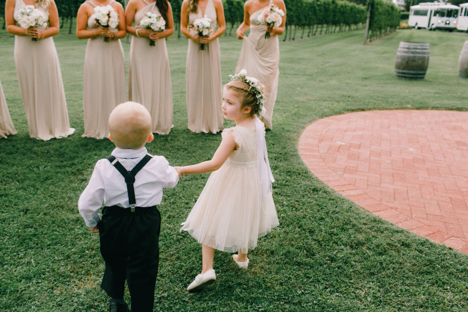 At the Willow Creek Winery Wedding, a young boy and girl in formal attire walk hand in hand on the lush grass, surrounded by bridesmaids in light dresses holding delicate bouquets.