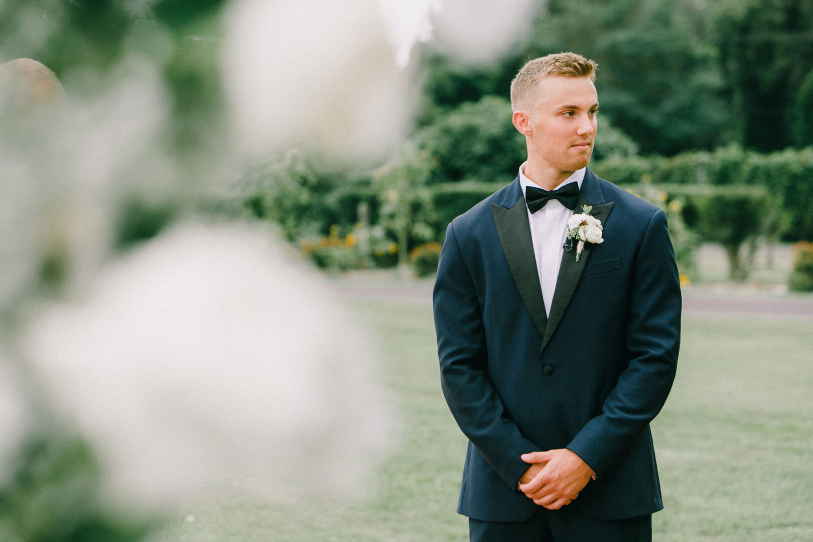 A man in a tuxedo stands outdoors on the grass at Willow Creek Winery, with trees and shrubs creating a picturesque wedding backdrop.