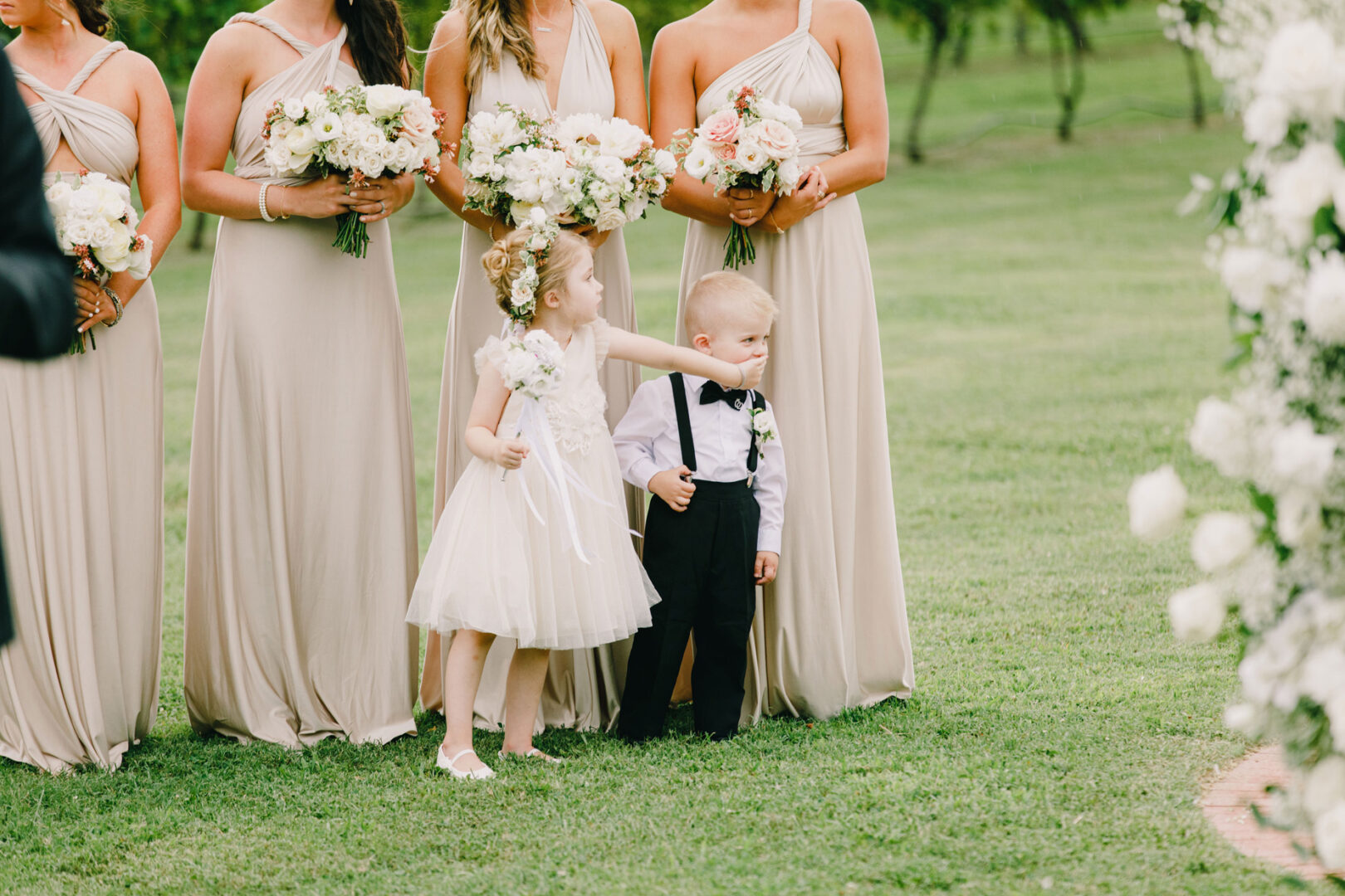 At a Willow Creek Winery wedding, a young girl in a white dress gently touches the face of a boy in formal attire, surrounded by adults holding flower bouquets in the garden setting.