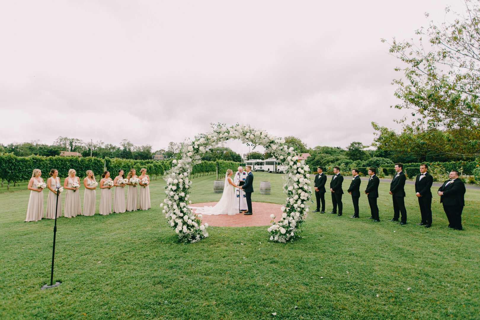 At the enchanting Willow Creek Winery wedding, a couple stands under a large floral arch, embraced by the vineyard's serene charm. Bridesmaids in beige dresses and groomsmen in black suits stand gracefully on either side, framing the picturesque outdoor ceremony.