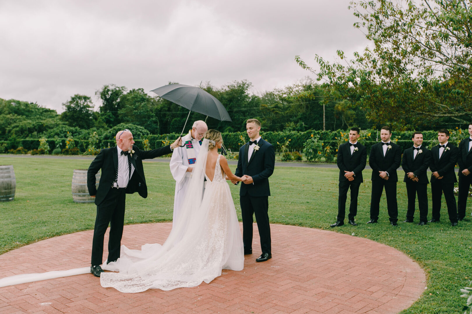 At the enchanting Willow Creek Winery wedding, a bride and groom stand elegantly in an outdoor ceremony. A man with an umbrella assists the officiant, while groomsmen pose gracefully in the background, adding to the idyllic scene.