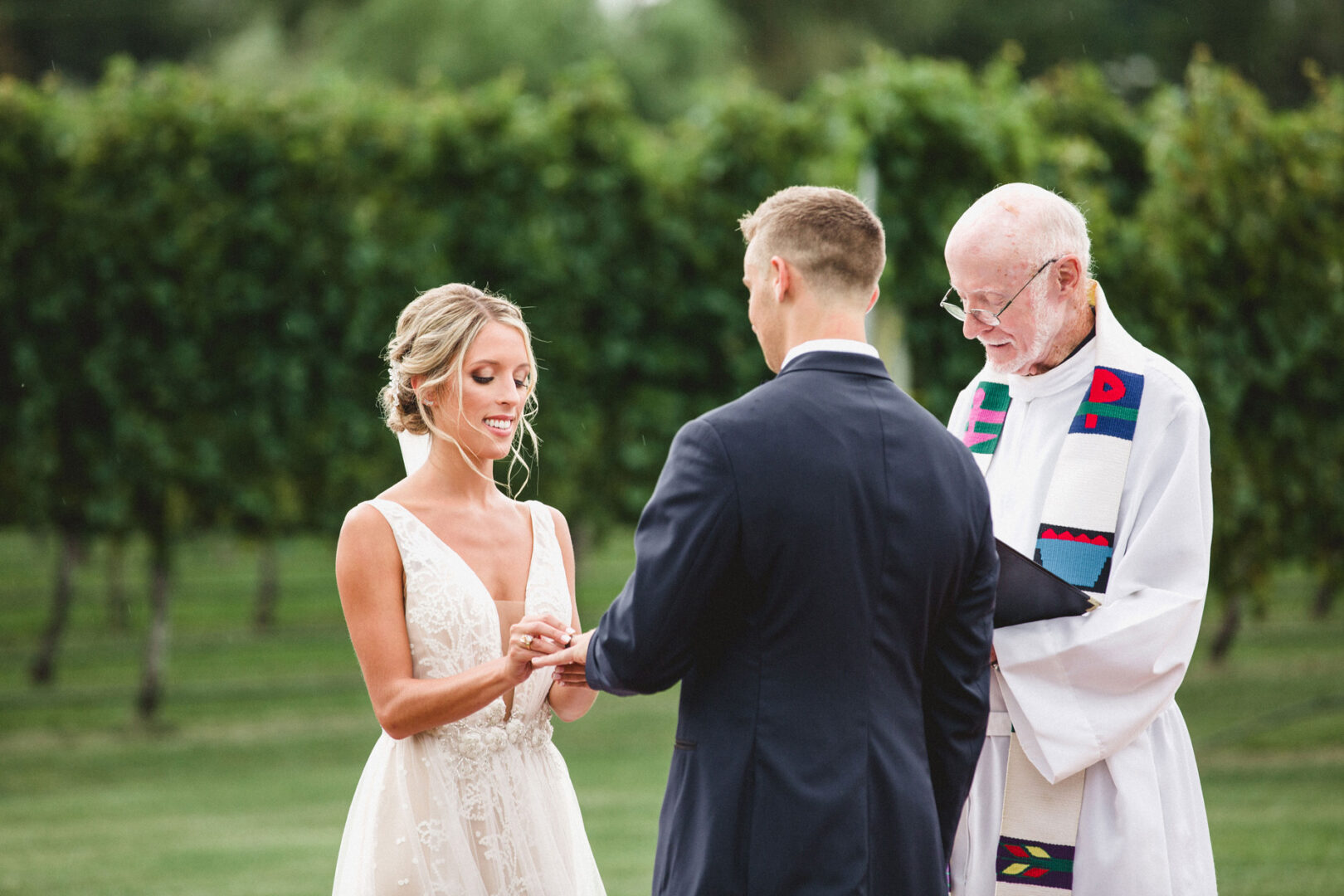 At Willow Creek Winery, a bride and groom exchange rings during an outdoor wedding ceremony officiated by a man in a white robe.