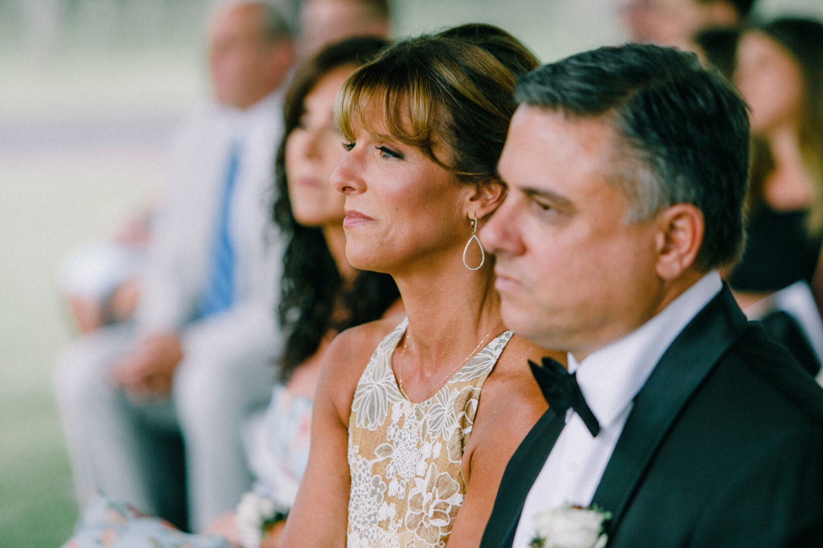At the Willow Creek Winery wedding, a woman and a man in formal attire sit outdoors among other guests, focusing intently on the event.