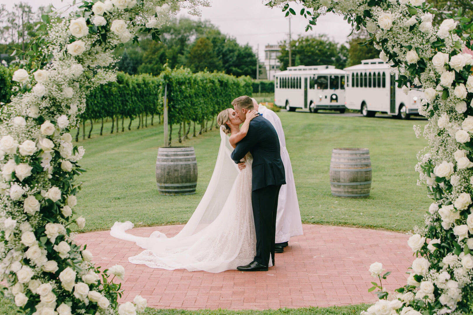 A bride and groom share a kiss under a floral arch at their Willow Creek Winery wedding. The picturesque scene is completed by white trolleys and lush vineyards in the background.