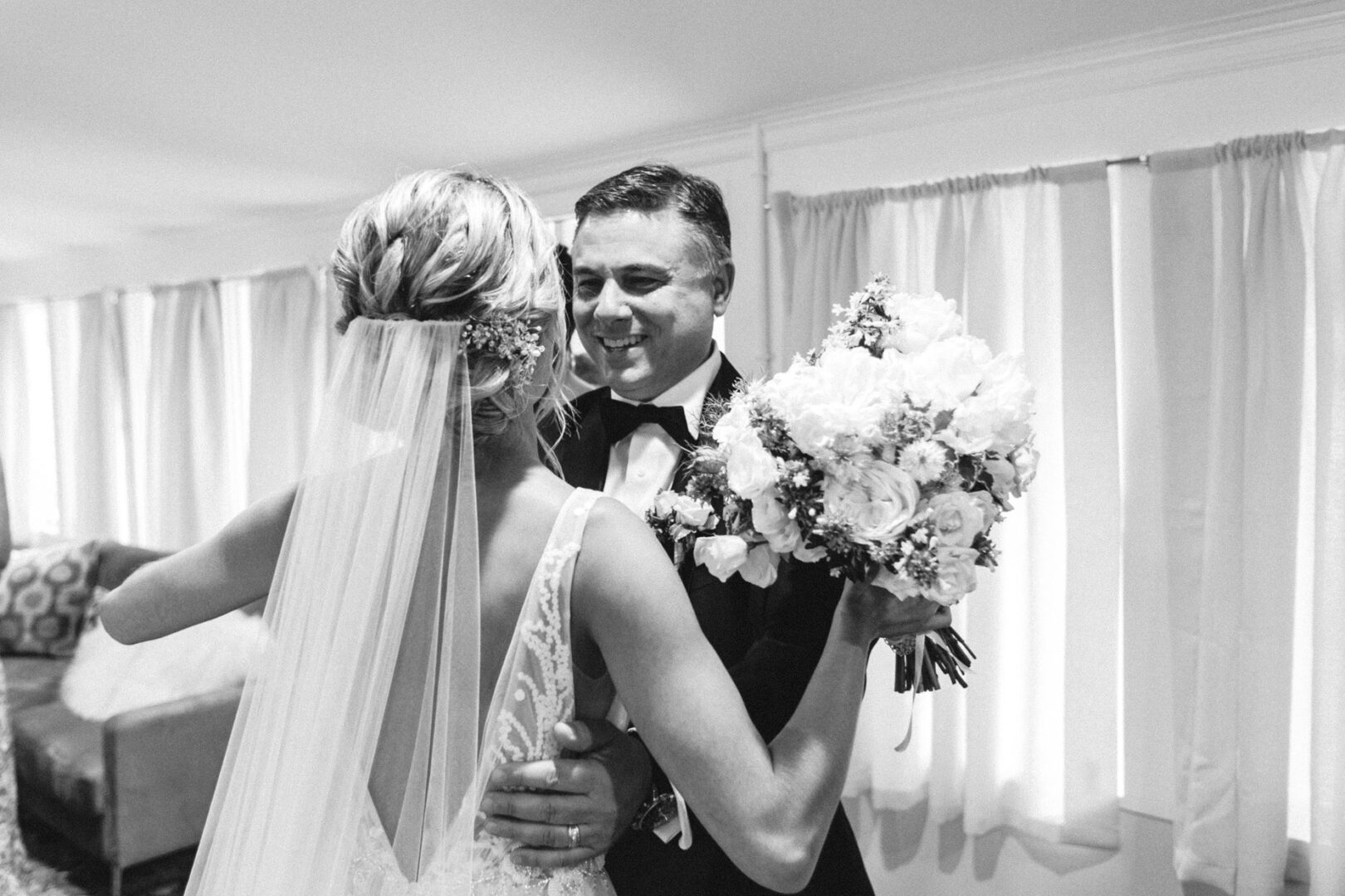 The bride and groom smile warmly at each other, the bride elegantly holding a bouquet, as soft curtains frame their moment in the enchanting ambiance of a Willow Creek Winery wedding.