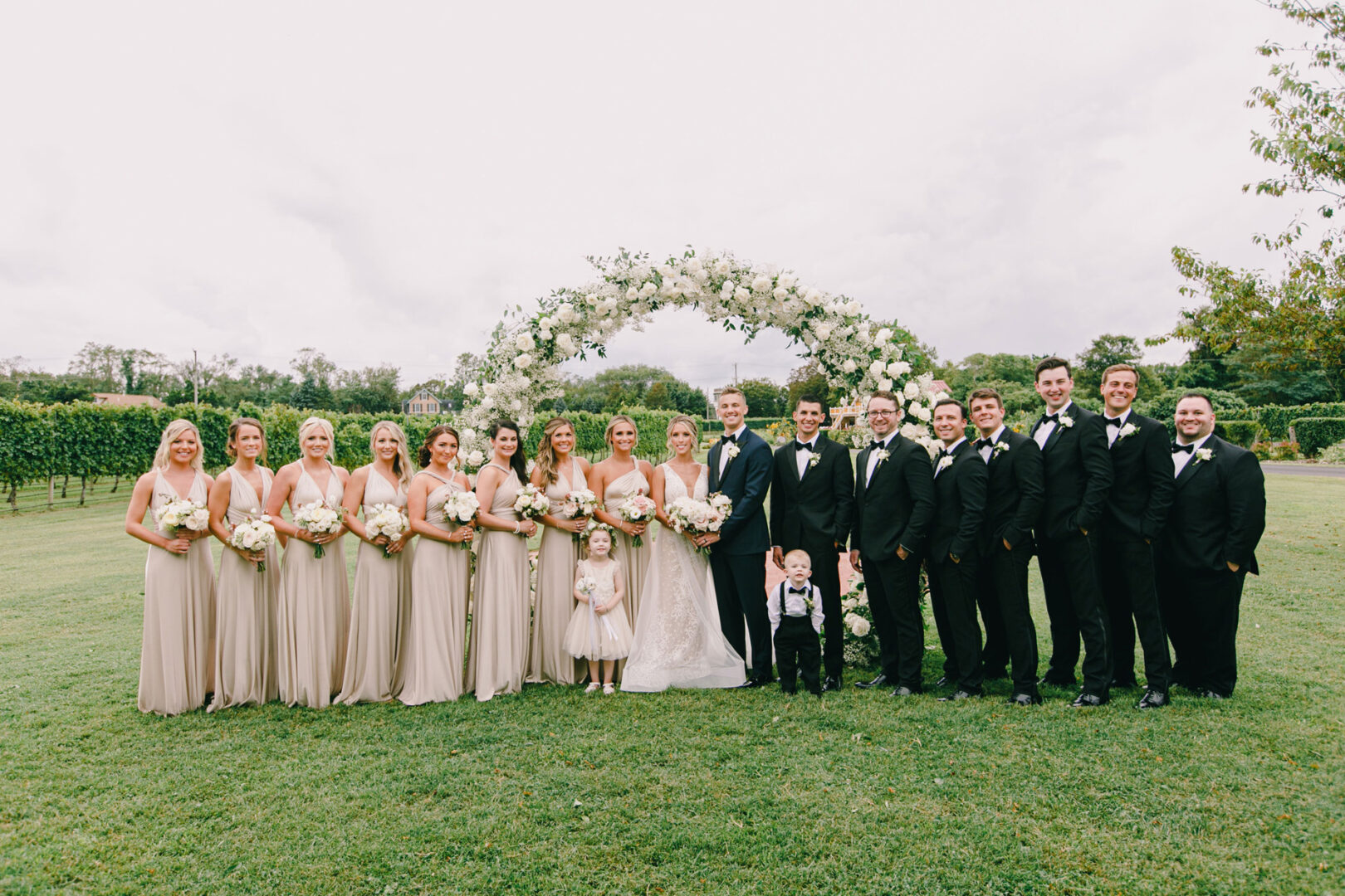 A wedding party poses on the lawn at Willow Creek Winery, featuring bridesmaids in beige dresses, groomsmen in black suits, flower girls, and ring bearers with a floral arch in the background.