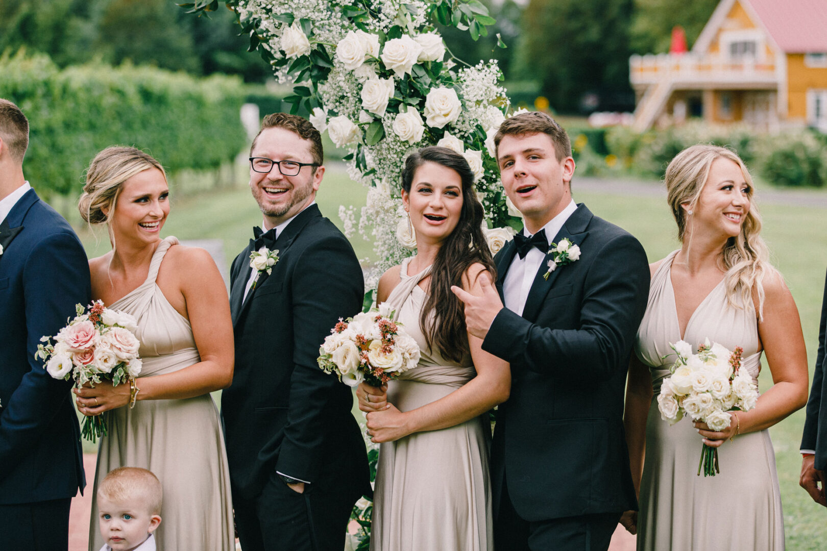 A group of five adults in formal attire stand together, smiling, with bouquets of flowers. A child is in the foreground at this Willow Creek Winery wedding. They are outdoors near a floral arch and greenery, creating a picturesque setting for the memorable event.