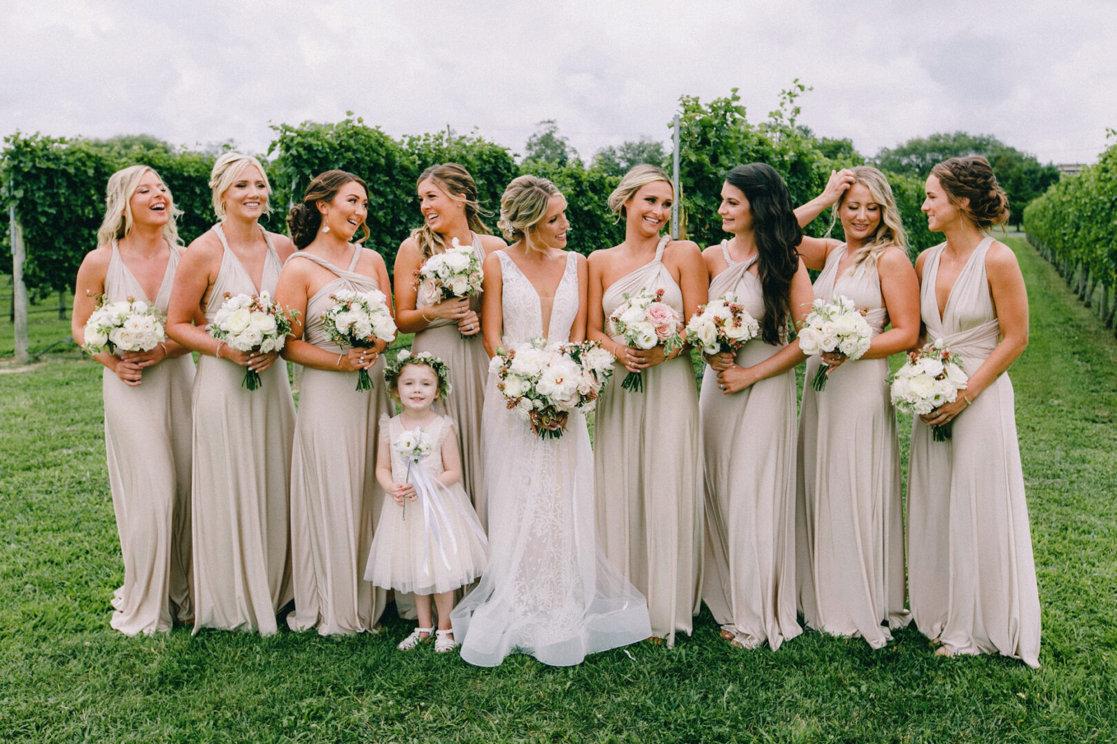 At Willow Creek Winery, a bride and eight bridesmaids line up outdoors with bouquets in hand. A young girl in a white dress stands before them, perfectly capturing the vineyard's enchanting setting.