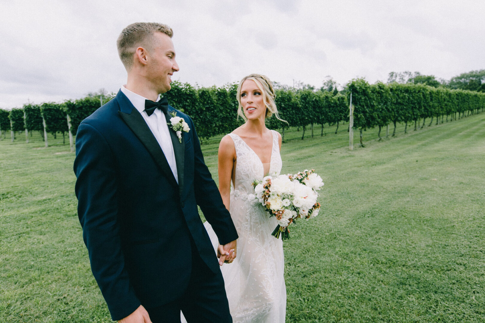 A bride and groom walk hand in hand through Willow Creek Winery's vineyard. The bride holds a bouquet of white flowers, and the groom is in a suit. The sky is cloudy, casting an intimate ambiance over this enchanting winery wedding.
