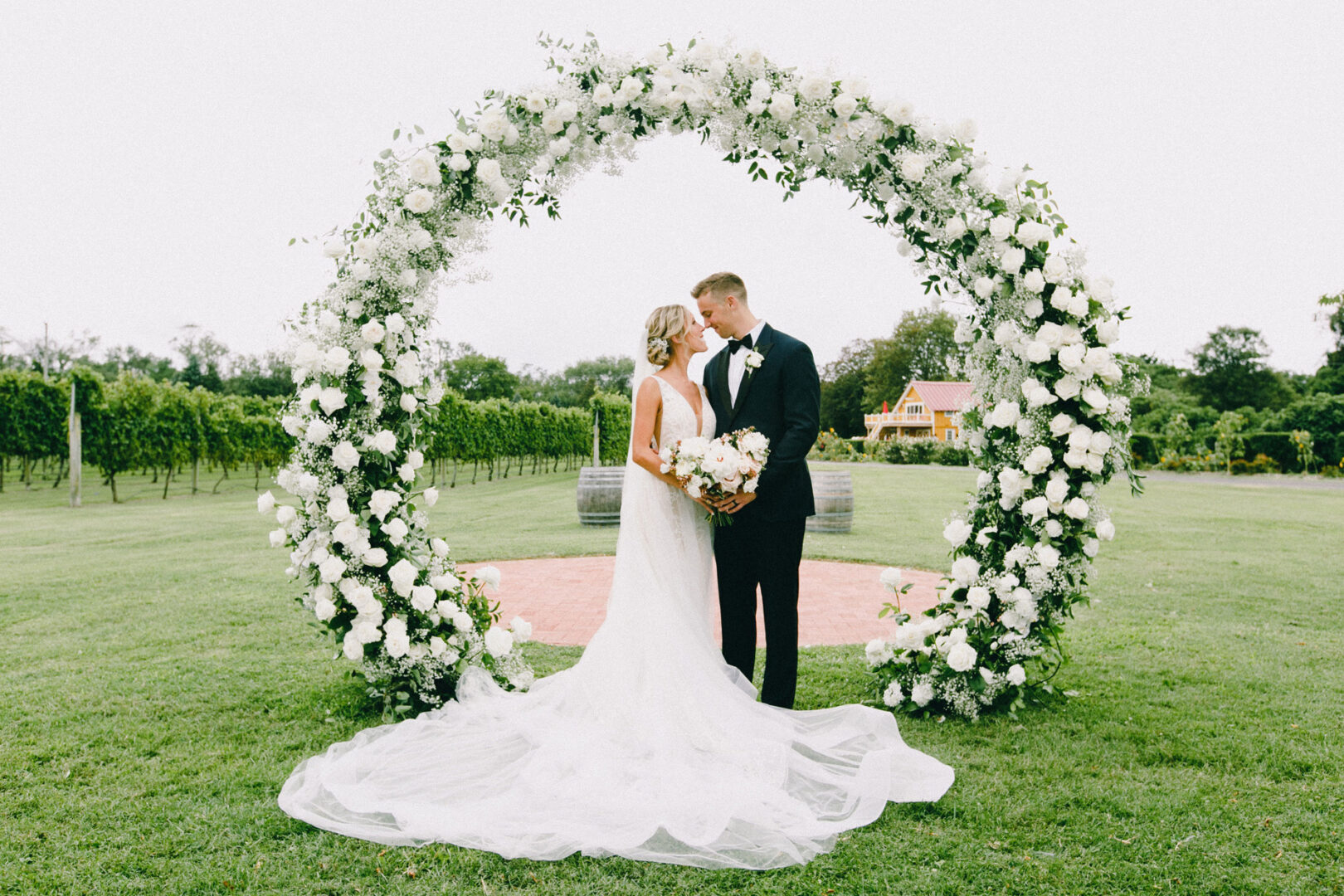 At Willow Creek Winery, a bride and groom stand facing each other under a large circular floral arch, surrounded by lush greenery in this enchanting outdoor setting.
