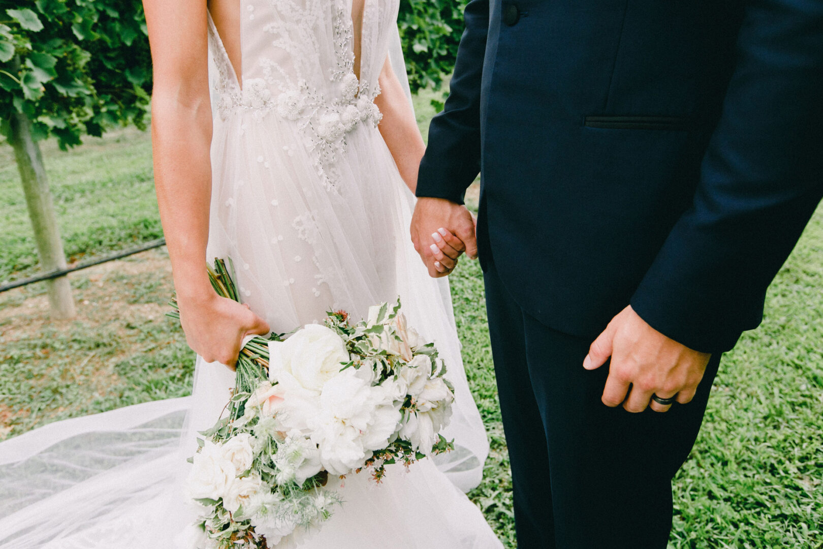A couple holds hands outdoors at Willow Creek Winery. The bride glows in a white dress with a bouquet of white flowers, while the groom looks dashing in a dark suit, both enjoying their enchanting wedding day.