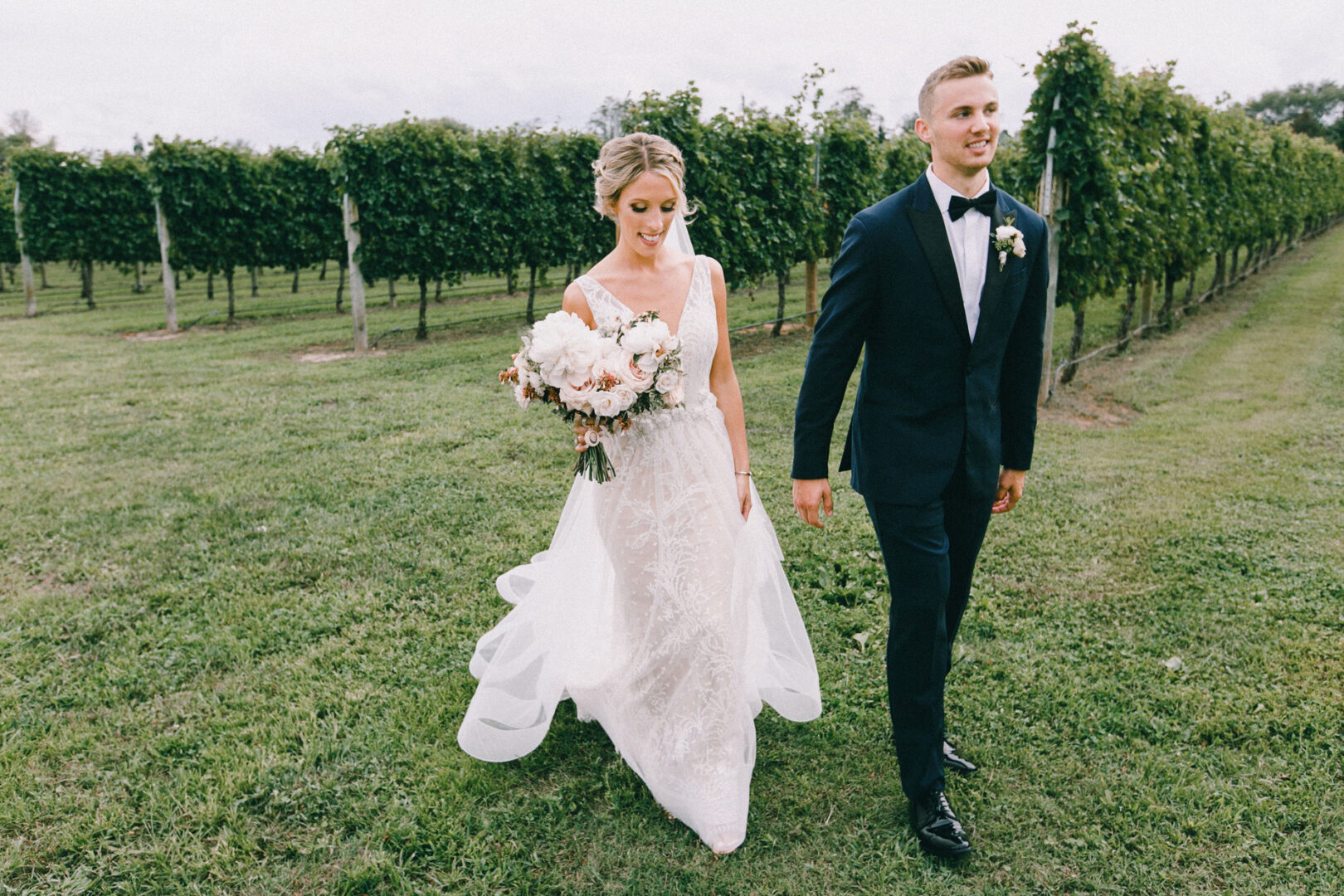 At the picturesque Willow Creek Winery, a bride and groom stroll side by side through the vineyard. The bride clutches a bouquet as they both exude elegance in their formal wedding attire.