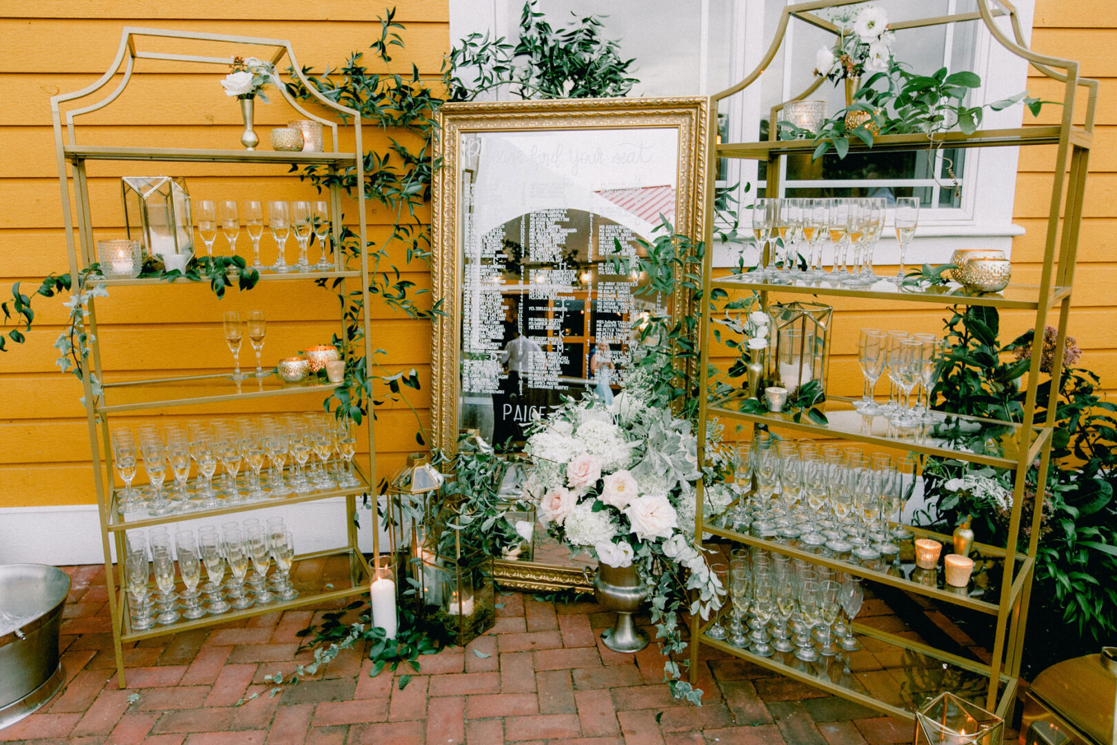 Experience the elegance of a Willow Creek Winery wedding with gold shelves adorned with glasses, a decorative mirror seating chart, and stunning floral arrangements set against a vibrant yellow wall.