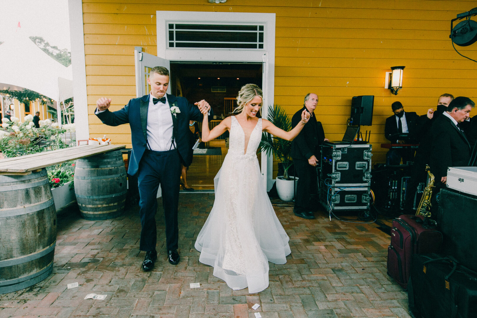 A bride and groom dance joyfully outside a yellow building at Willow Creek Winery, surrounded by musicians and their equipment.