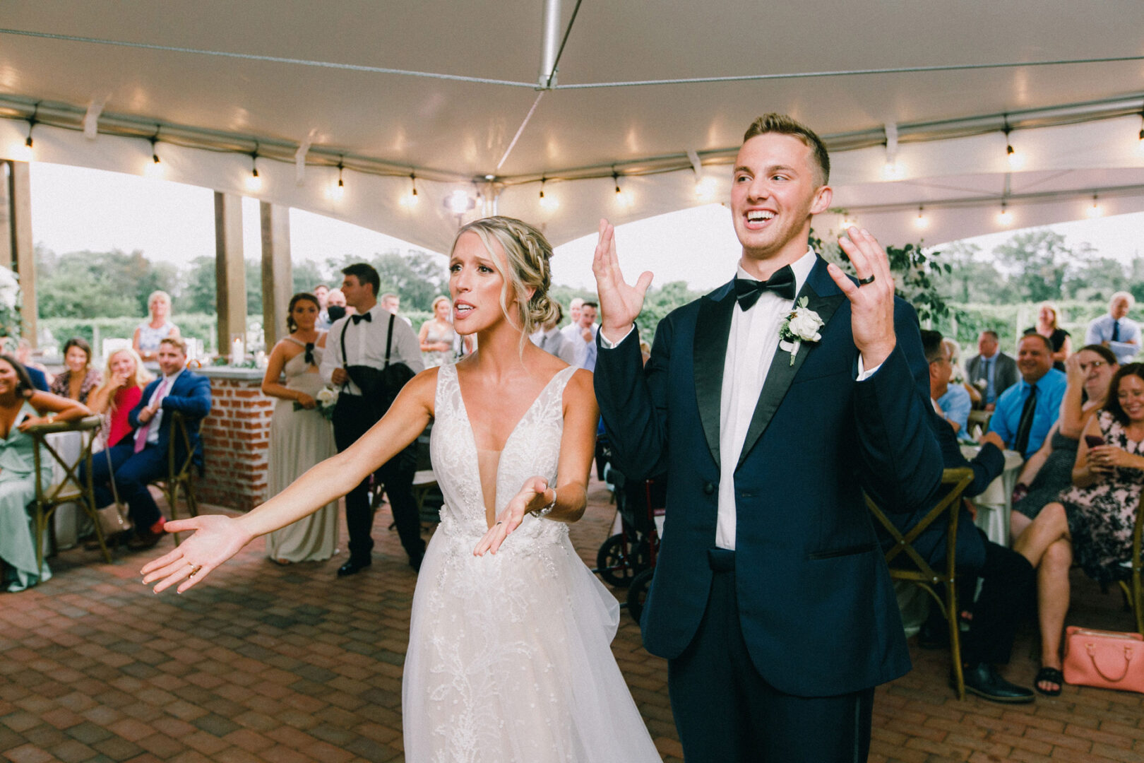 A bride and groom in formal attire gesture expressively under a tent at their Willow Creek Winery wedding reception, surrounded by seated guests.