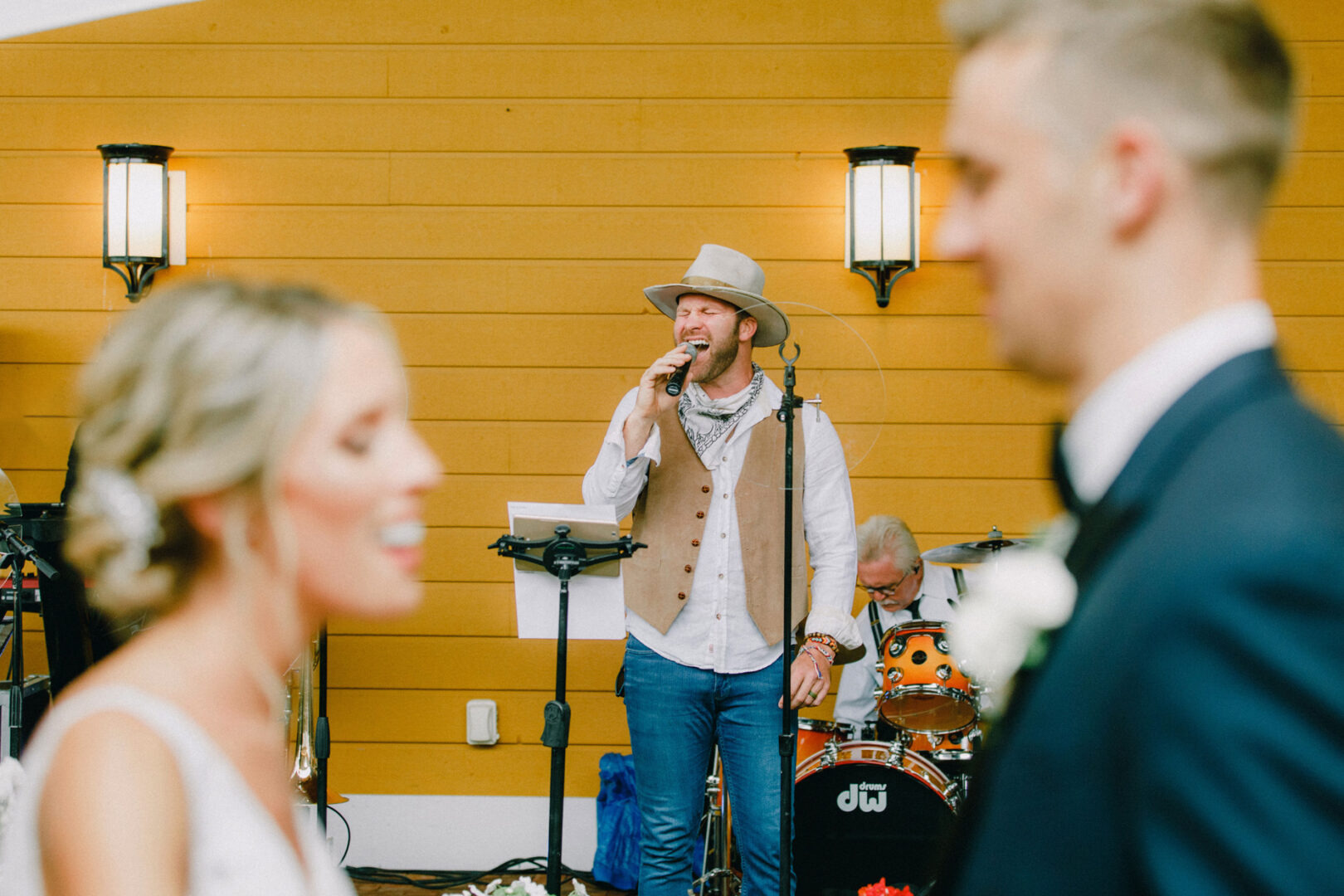 A singer with a hat performs on stage at the enchanting Willow Creek Winery Wedding, with a drummer behind, while a couple stands in the foreground.