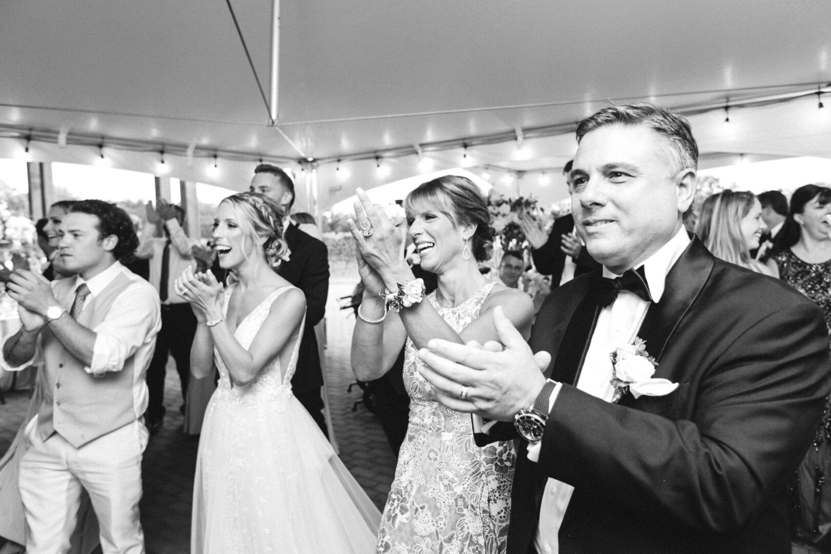 A group of people in formal attire cheer and clap at a Willow Creek Winery wedding, celebrating joyously under a tent.