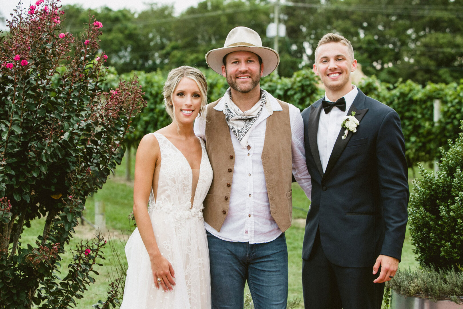 A bride in a white dress, a man in casual attire with a hat, and a groom in a suit stand together outdoors at the picturesque Willow Creek Winery, surrounded by lush greenery.