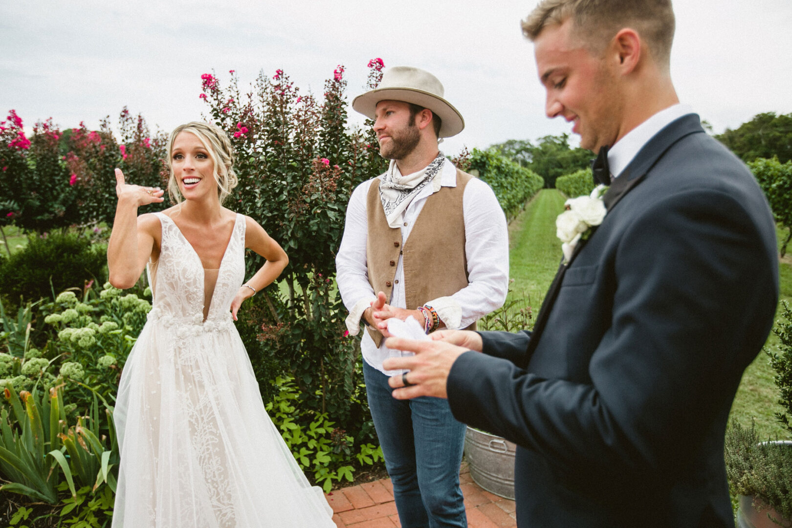 At the enchanting Willow Creek Winery wedding, a bride playfully gestures while standing next to a casually dressed man and a smiling groom in the lush garden setting.