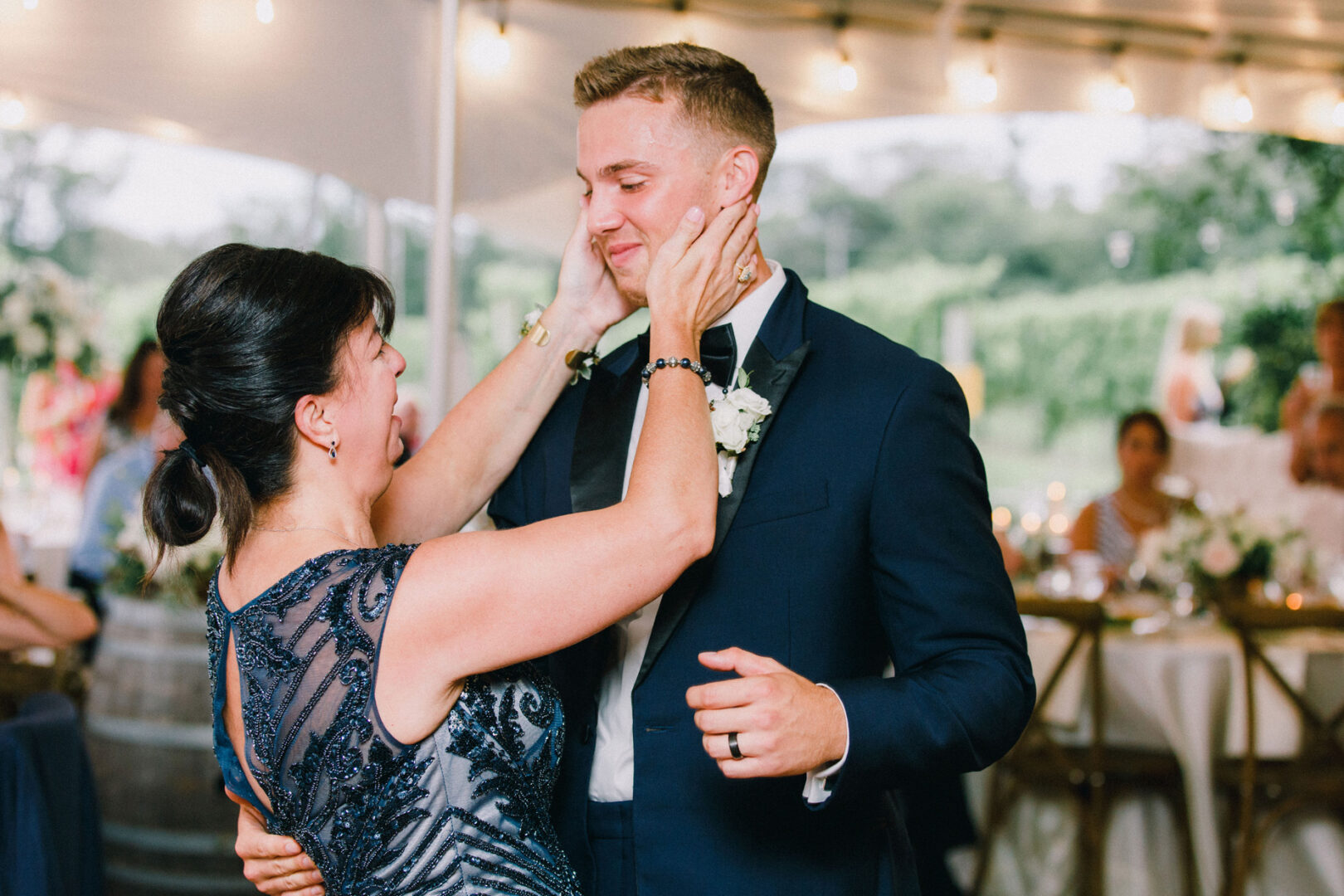 At a Willow Creek Winery wedding, a woman in a blue dress embraces a man in a suit with a boutonniere, both smiling warmly. They stand under a tent adorned with string lights, creating an enchanting atmosphere for the celebration.