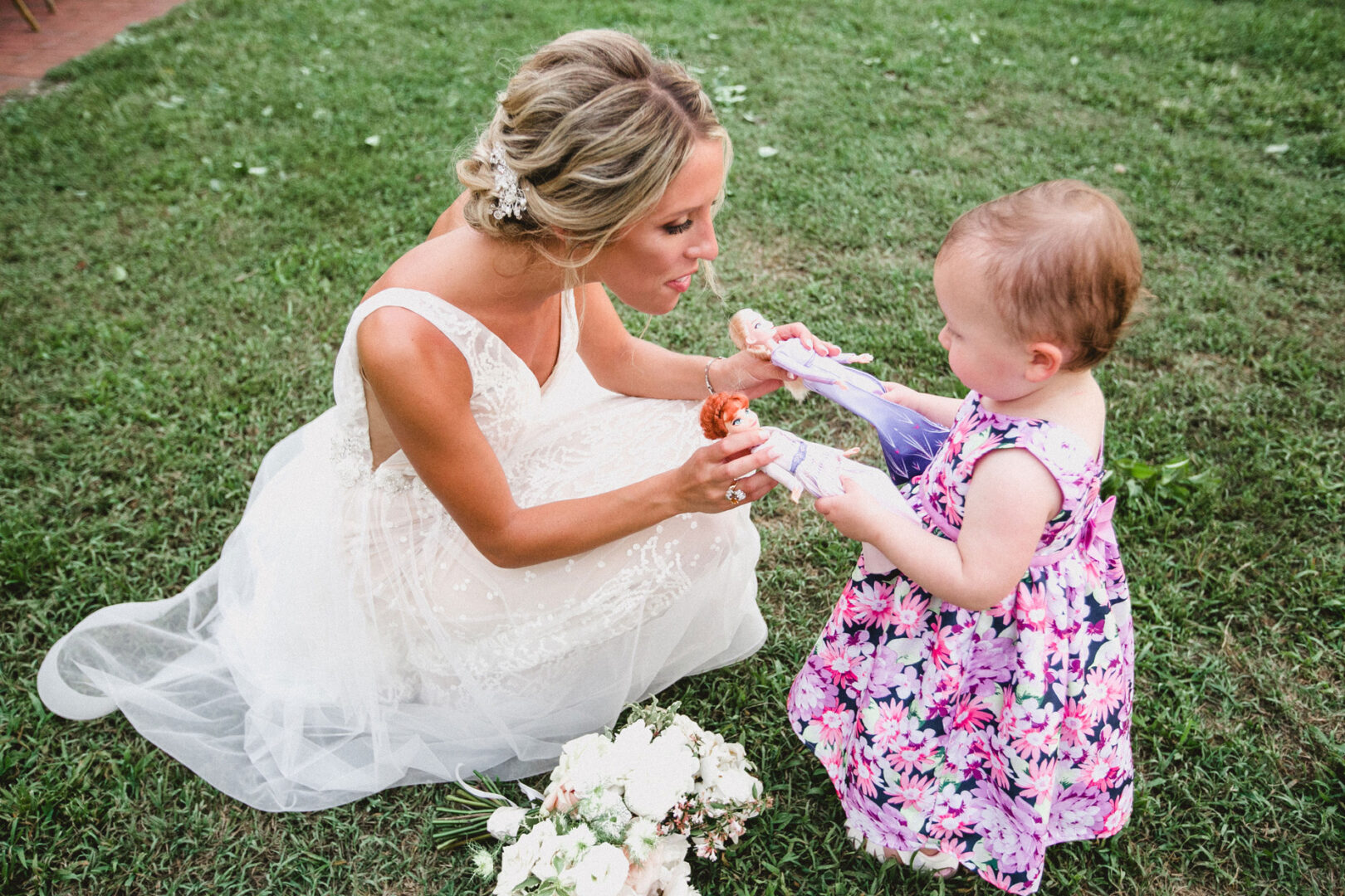 At a Willow Creek Winery wedding, a woman in a white dress kneels on the grass, engaging with a young girl in a floral dress holding a popsicle. Nearby, a bouquet lies elegantly.