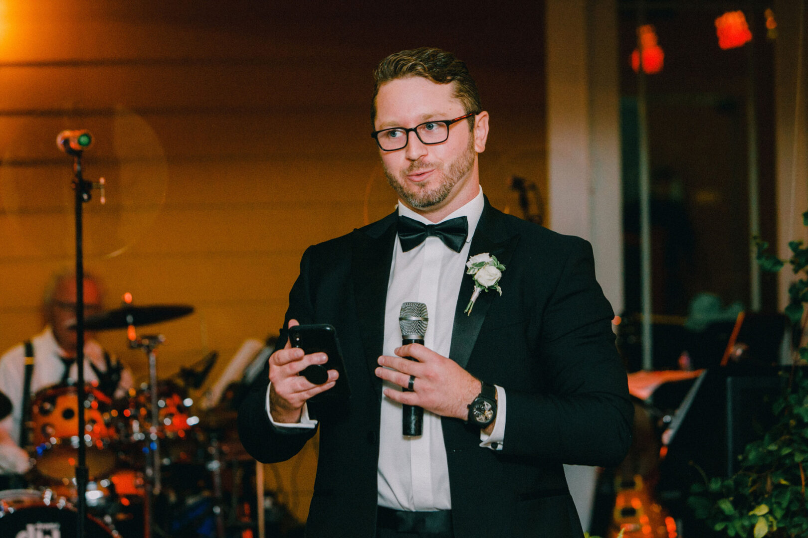 At the Willow Creek Winery wedding, a man in a tuxedo speaks into a microphone, holding a phone, with a band playing lively tunes in the background.