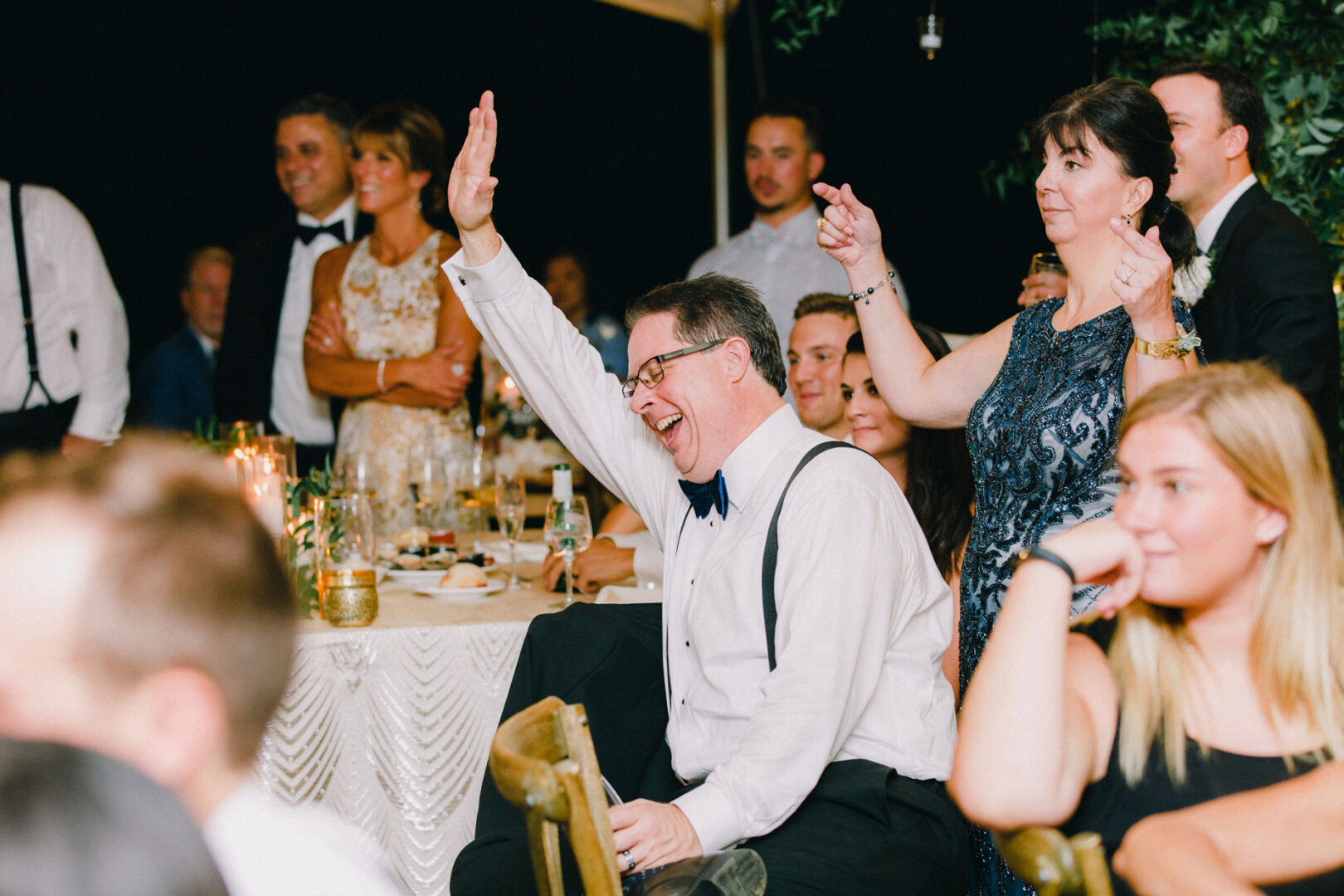 At a Willow Creek Winery wedding, a man in a white shirt and bow tie raises his hand, smiling warmly at the formal event. Guests around him are seated, with a woman pointing to him as others observe. The tables are elegantly adorned with candles and decorations.