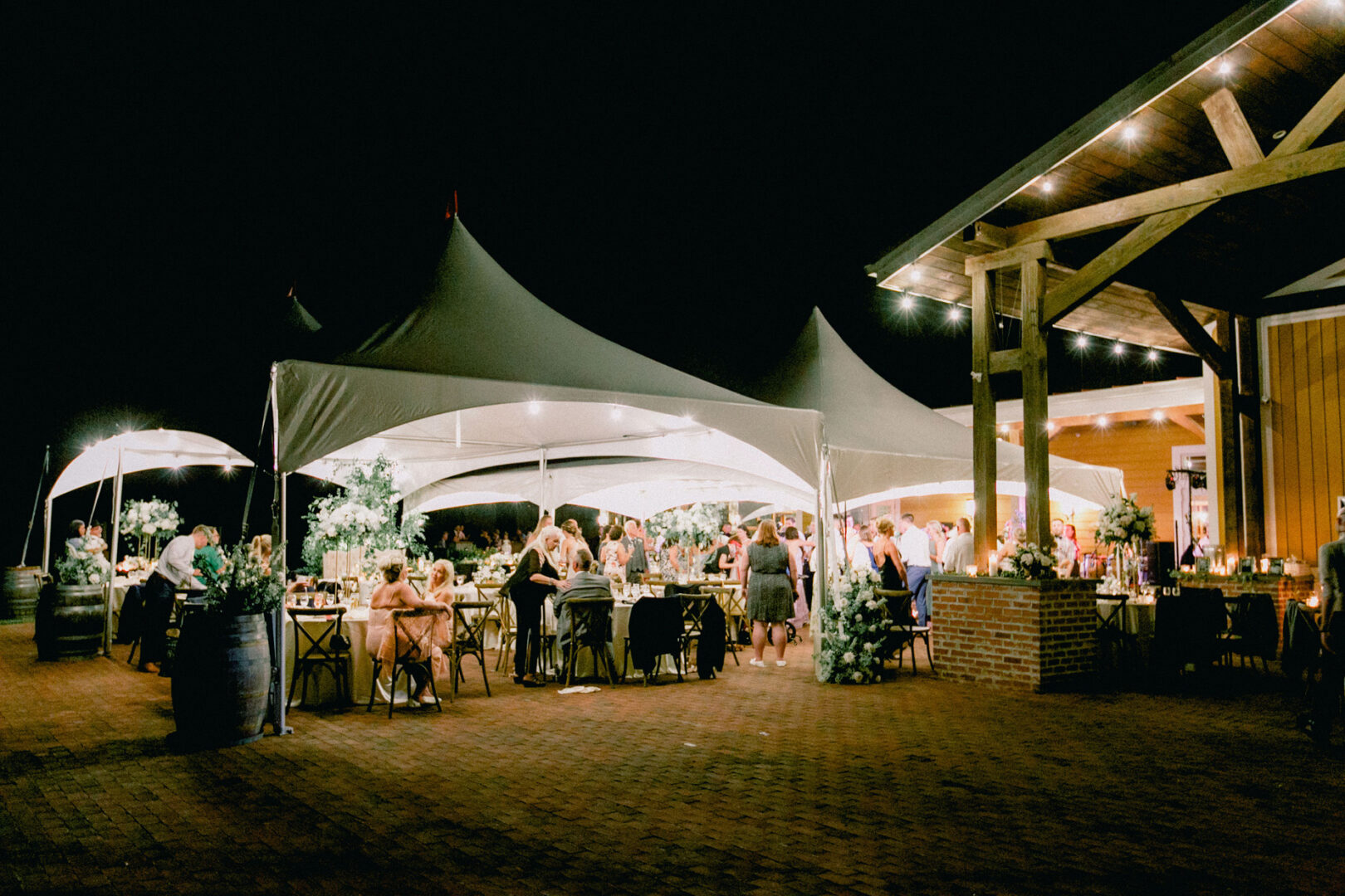The enchanting evening unfolded at Willow Creek Winery, where an outdoor event under large tents was adorned with string lights and lush greenery. Guests stood and sat amidst the idyllic setting, savoring the wedding's serene ambiance.