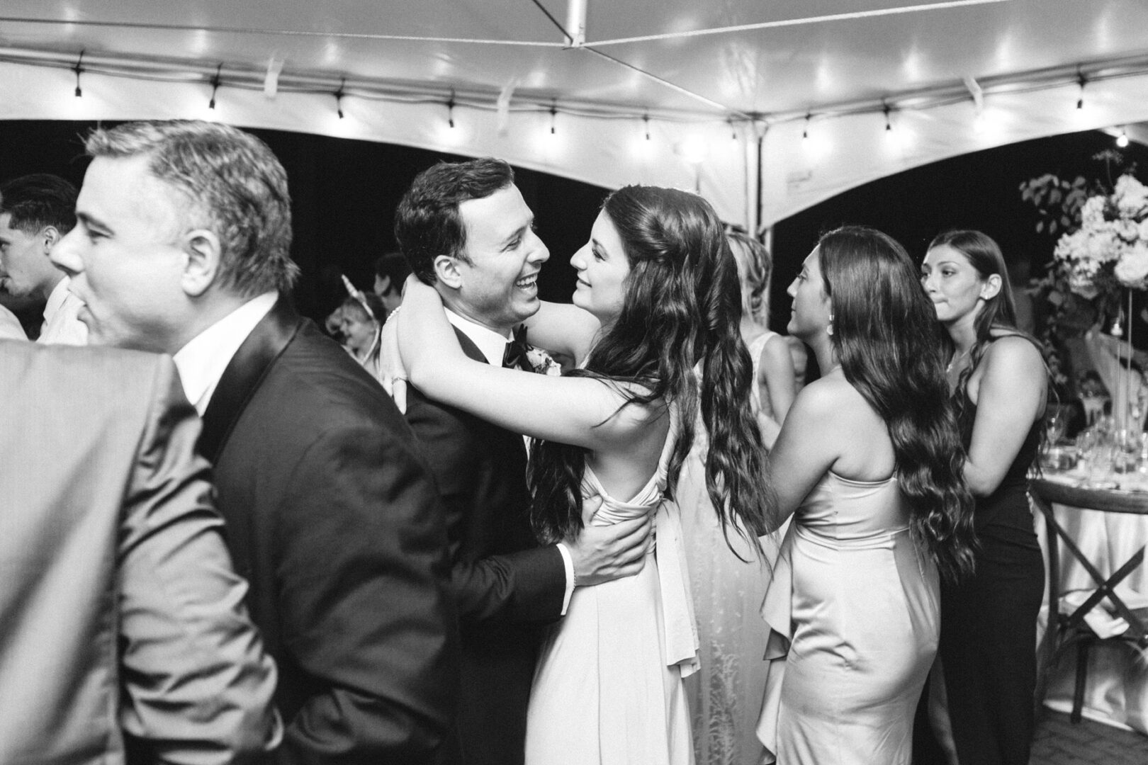 At a Willow Creek Winery wedding, a couple dressed formally embrace each other, surrounded by joyful guests under an elegant tent.