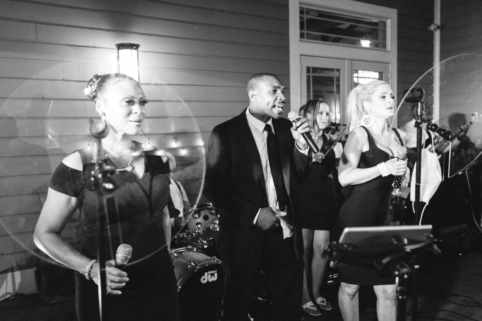 A black and white photo captures a band performing at a Willow Creek Winery wedding. Three singers with microphones are in focus, while a drummer and violinist add to the enchanting atmosphere in the background.