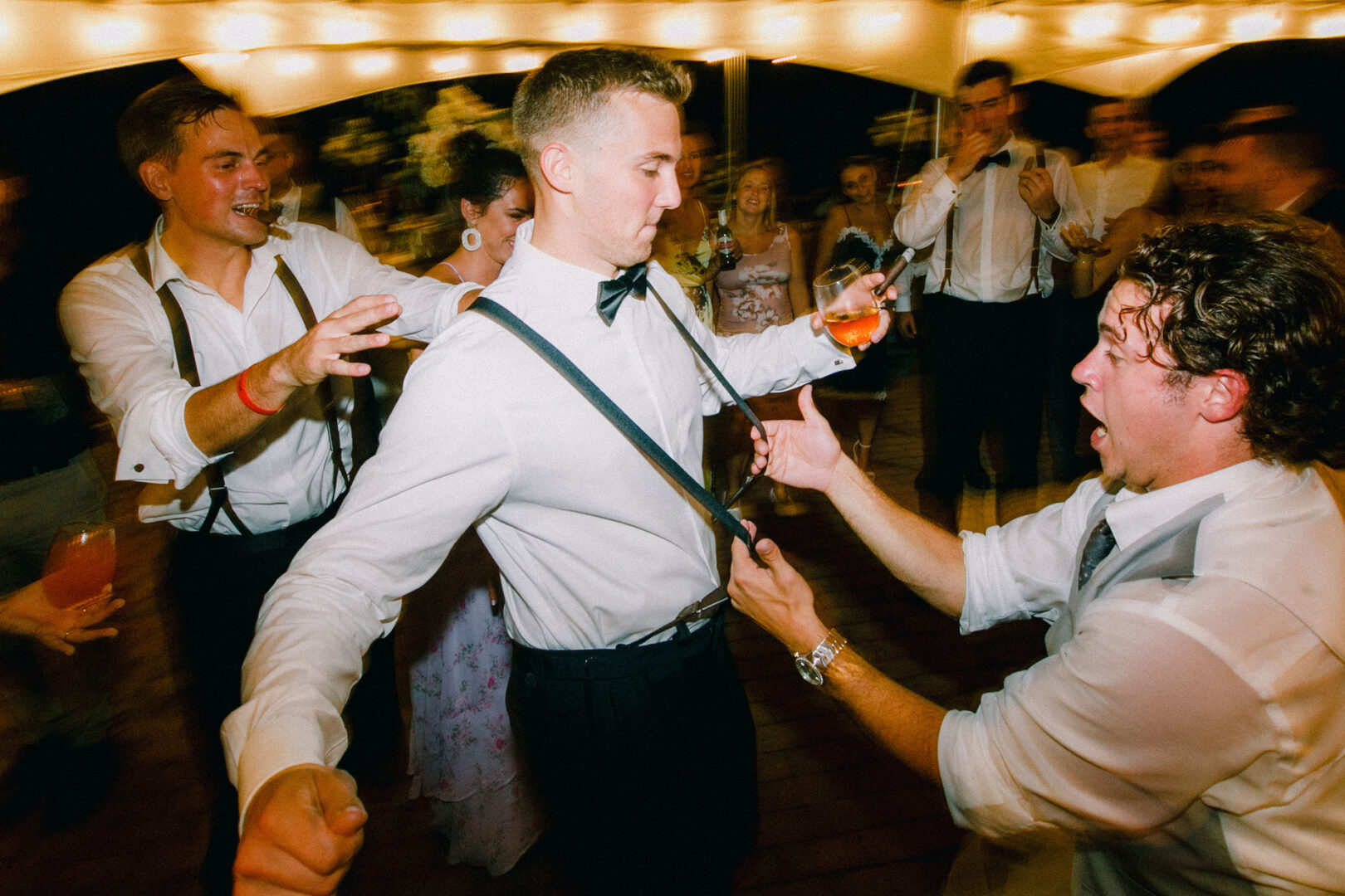 At a lively Willow Creek Winery wedding, a group of people dance energetically, with two men in the foreground playfully holding each other's suspenders, adding charm to the festive scene.